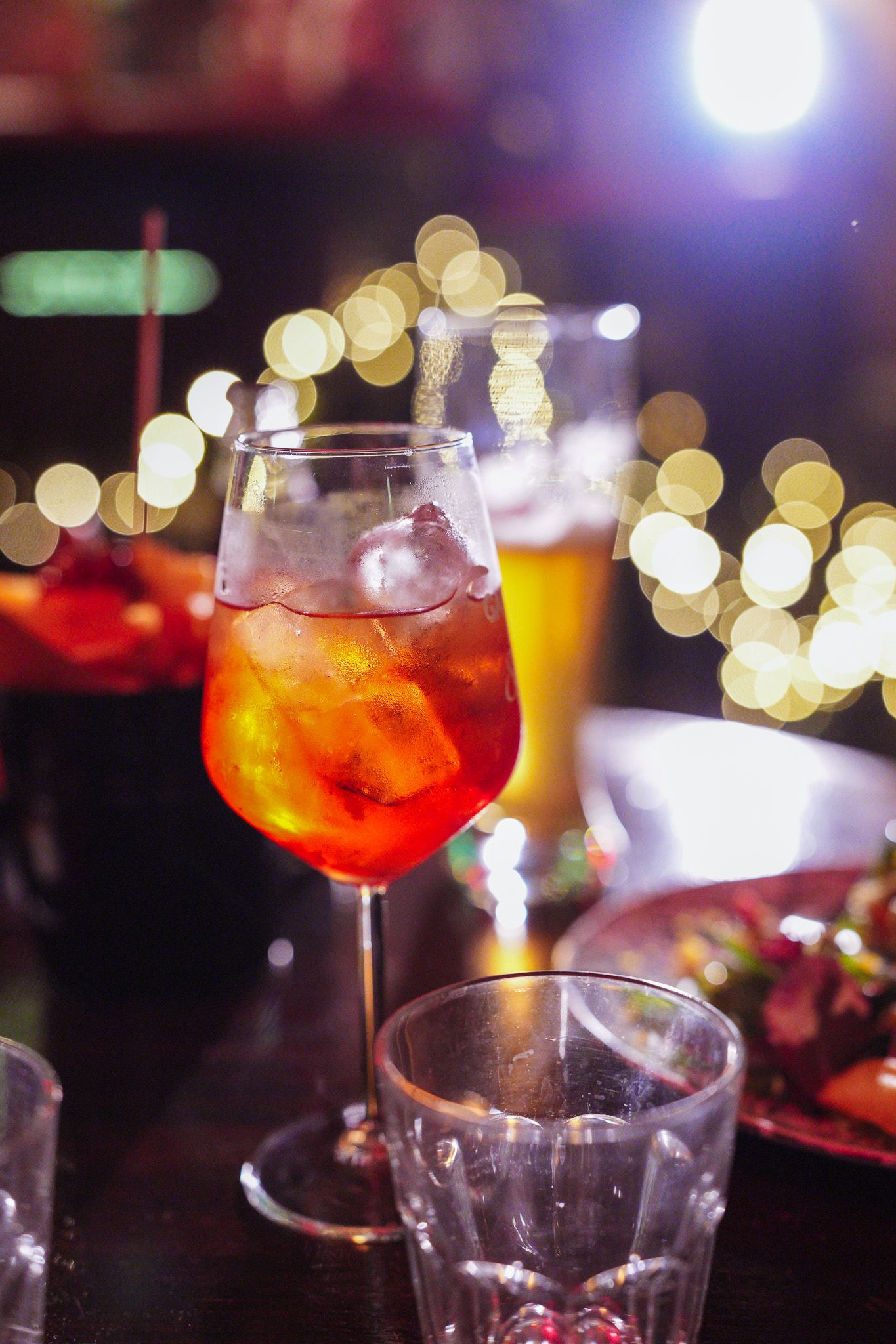 Cocktail con ghiaccio, una birra alta e uno stuzzichino su un tavolo, con lucine sullo sfondo.