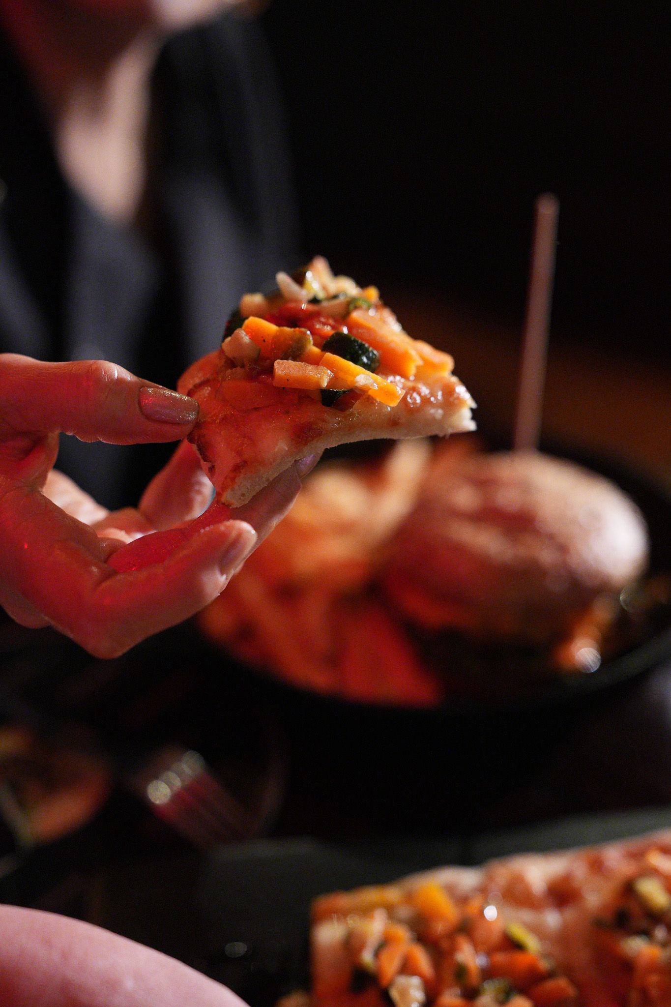 Mano che tiene una fetta di pizza con verdure, sullo sfondo sfocato altro cibo su un tavolo.