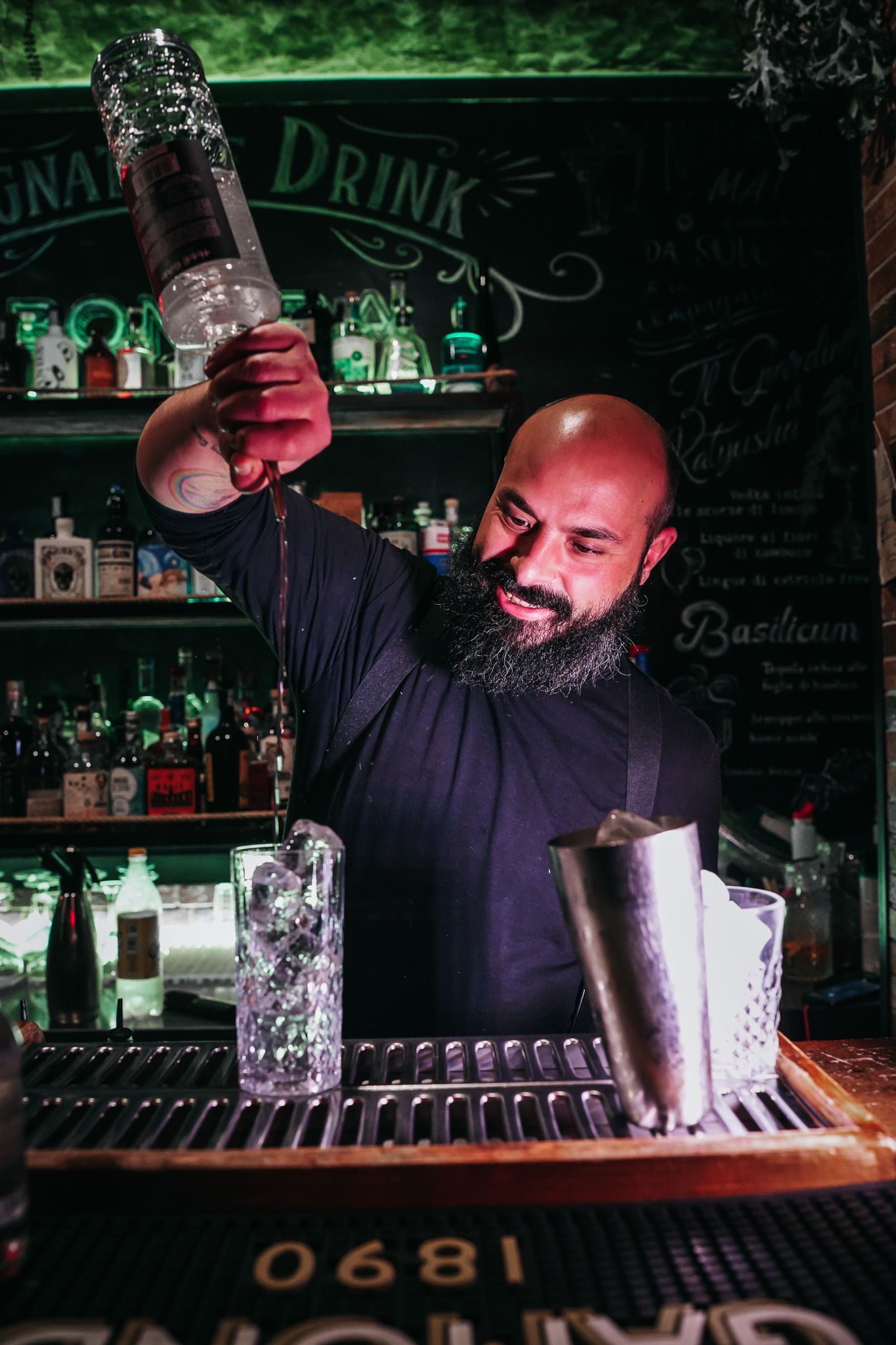 Barista che versa un liquido trasparente in un bicchiere con ghiaccio, dietro al bancone.