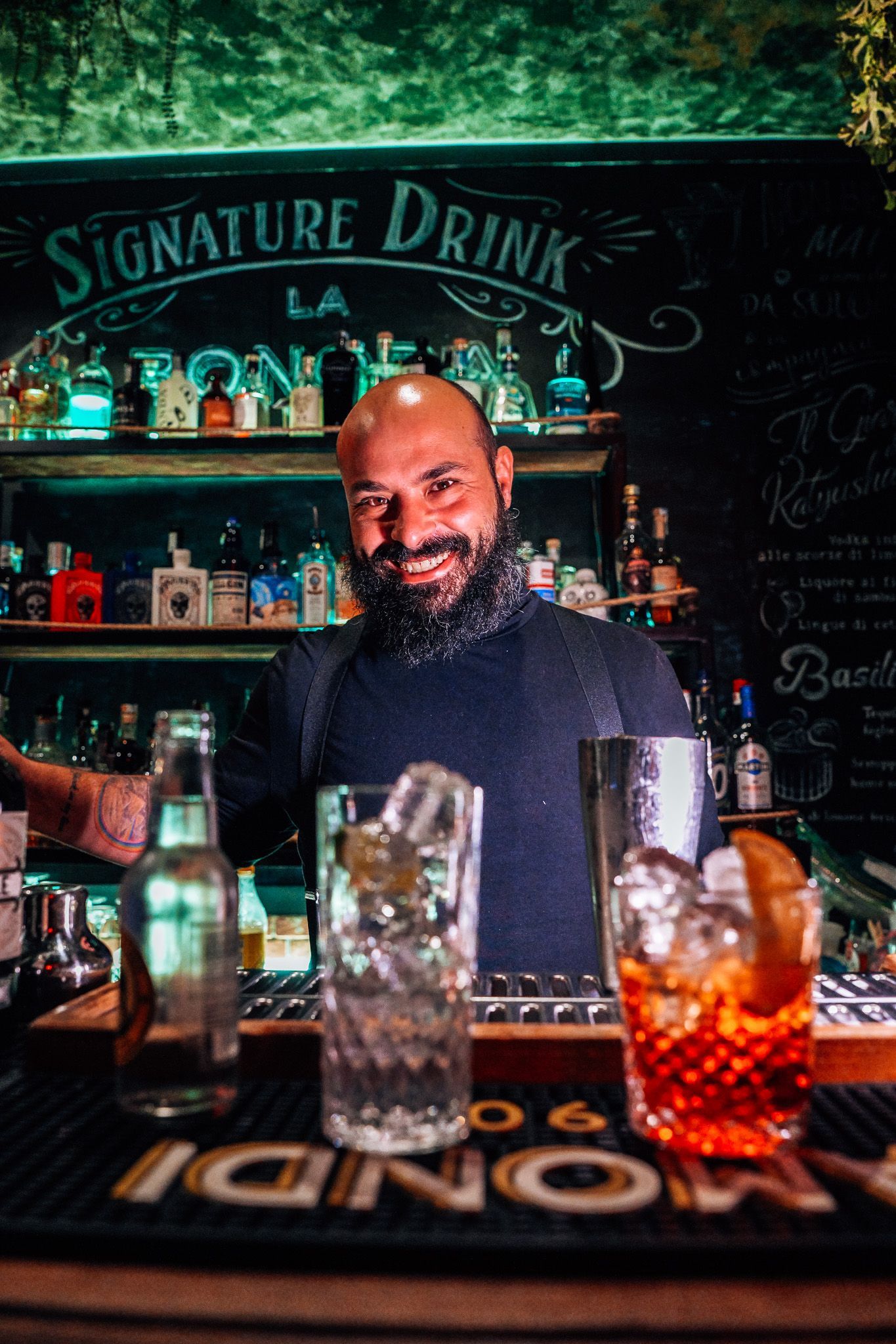 Barista sorridente al bancone, con due cocktail e bottiglie di liquore sullo sfondo.