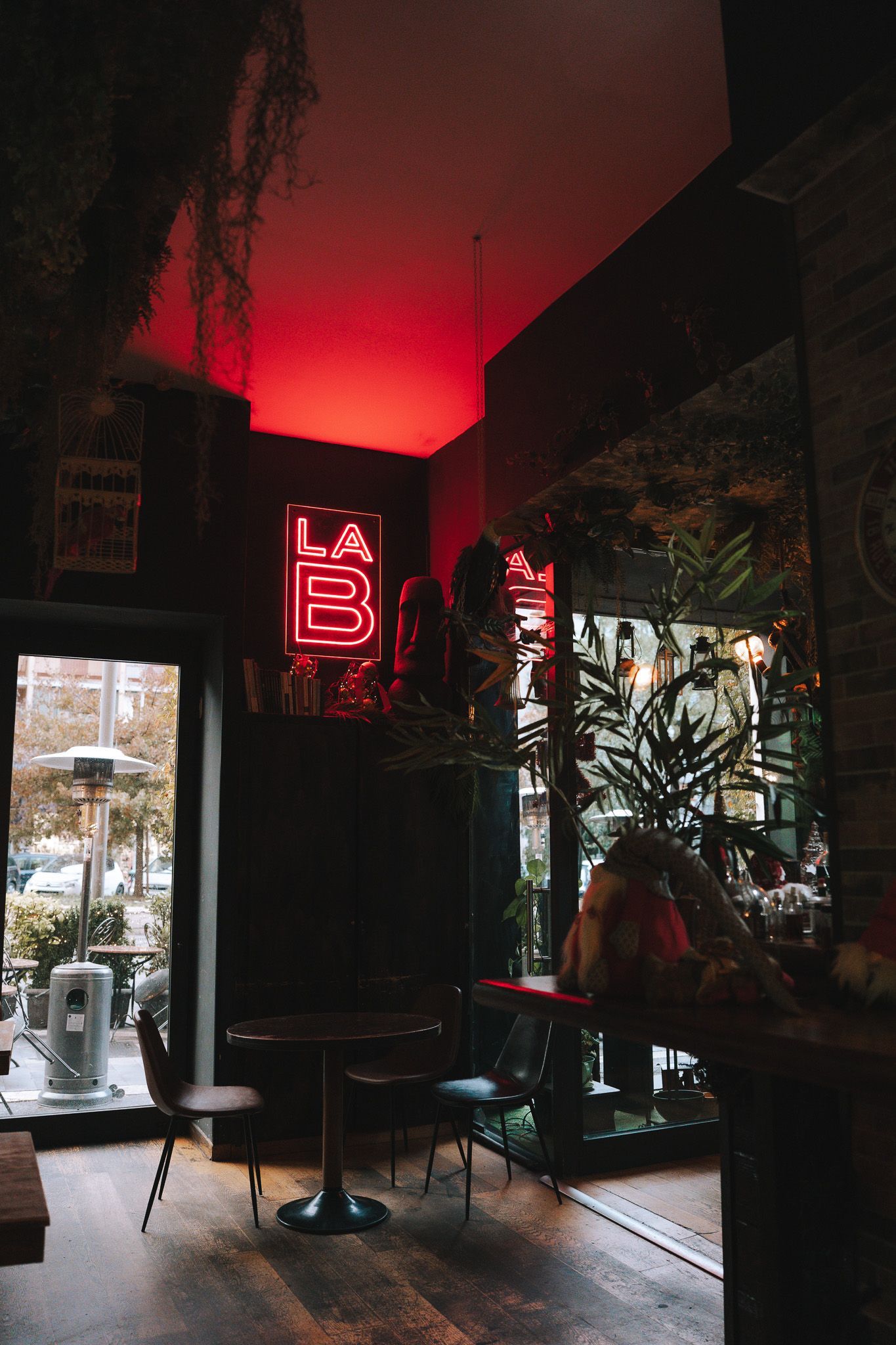 Interno scuro con insegna al neon rossa, tavolini e vetrata con piante, vista sull'esterno.