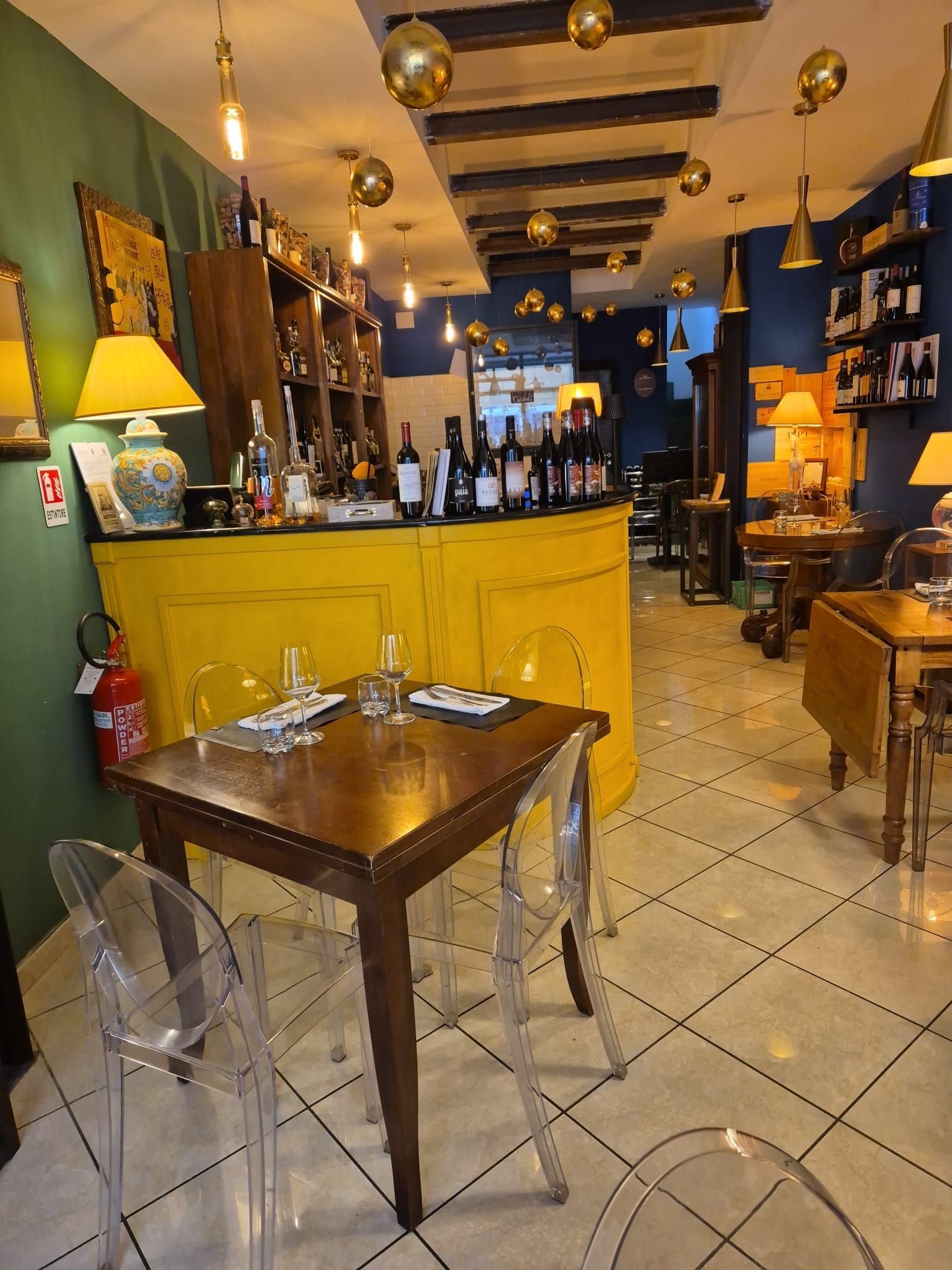 Interno di un ristorante con bancone giallo, tavoli in legno, sedie trasparenti, bottiglie di vino e luci sospese.