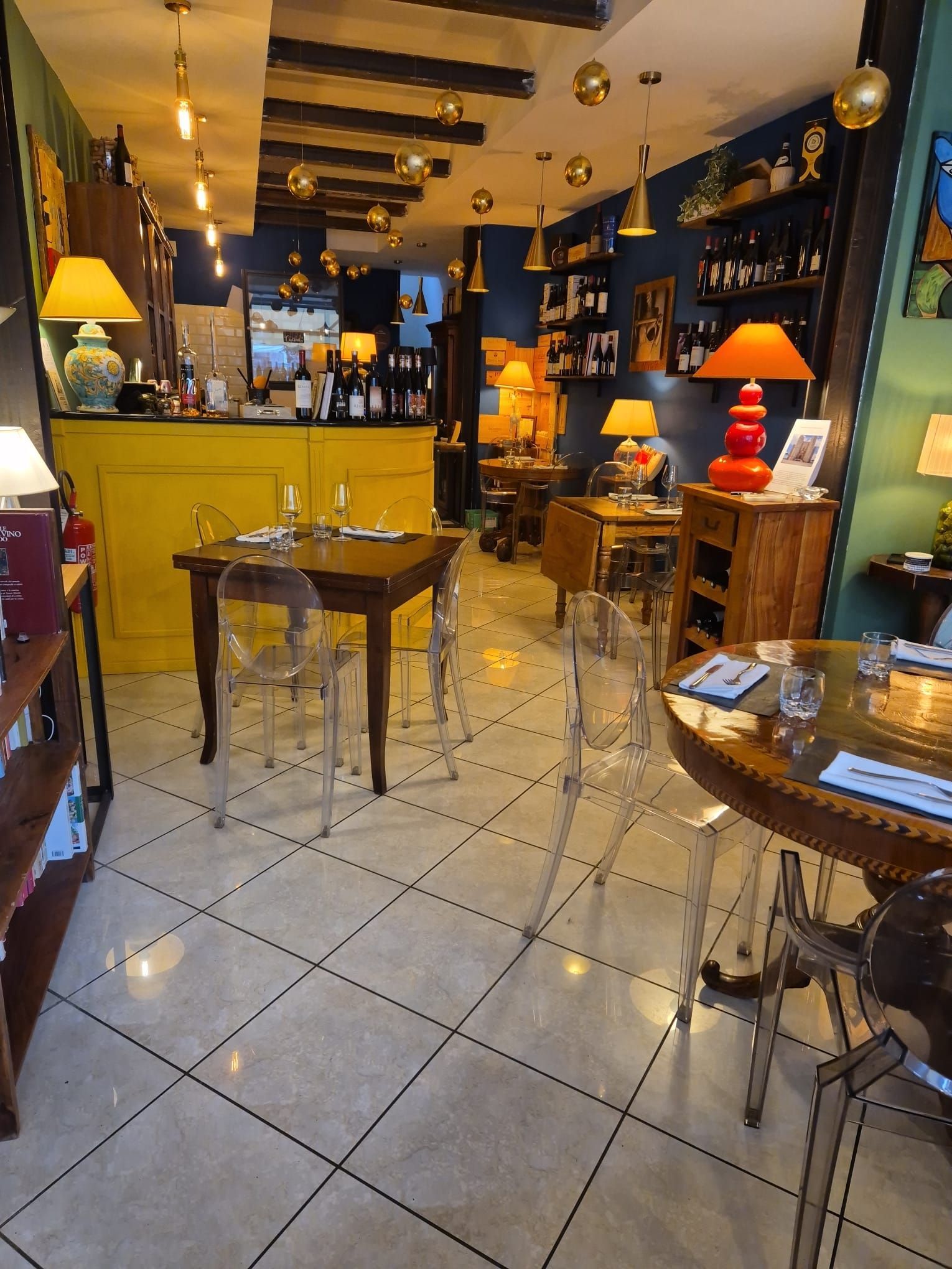 Interno del ristorante con bancone giallo, tavoli, sedie, lampade e bottiglie di vino.