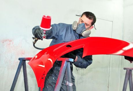 A man wearing a mask is spray painting a red bumper.