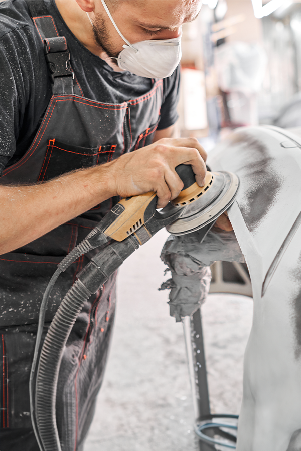 A man wearing a mask is sanding a car with a sander.