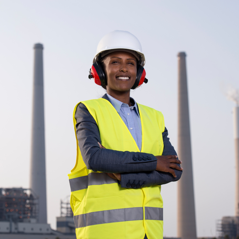 A man wearing a hard hat and headphones is standing in front of a factory