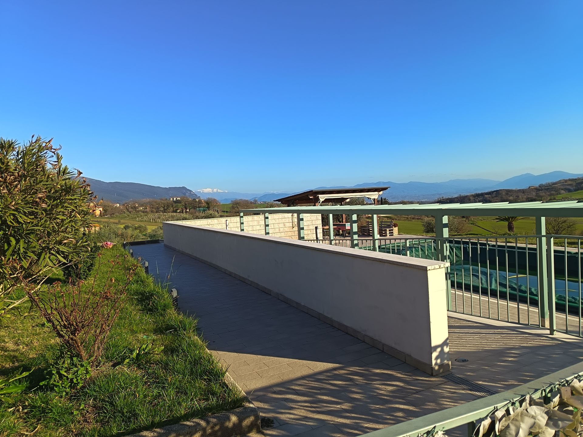 Un lungo muro beige costeggia un sentiero lastricato accanto a una struttura con cornice verde, affacciata su un paesaggio