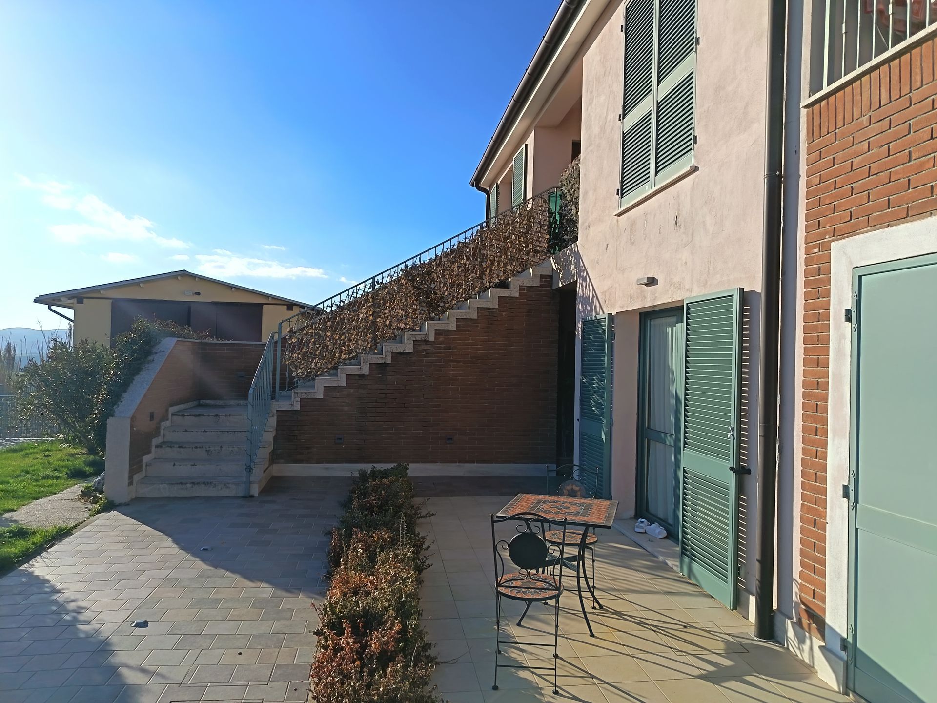 Un patio esterno in pietra con un set da bistrot e una scala esterna che conduce a un edificio a più piani di colore chiaro.