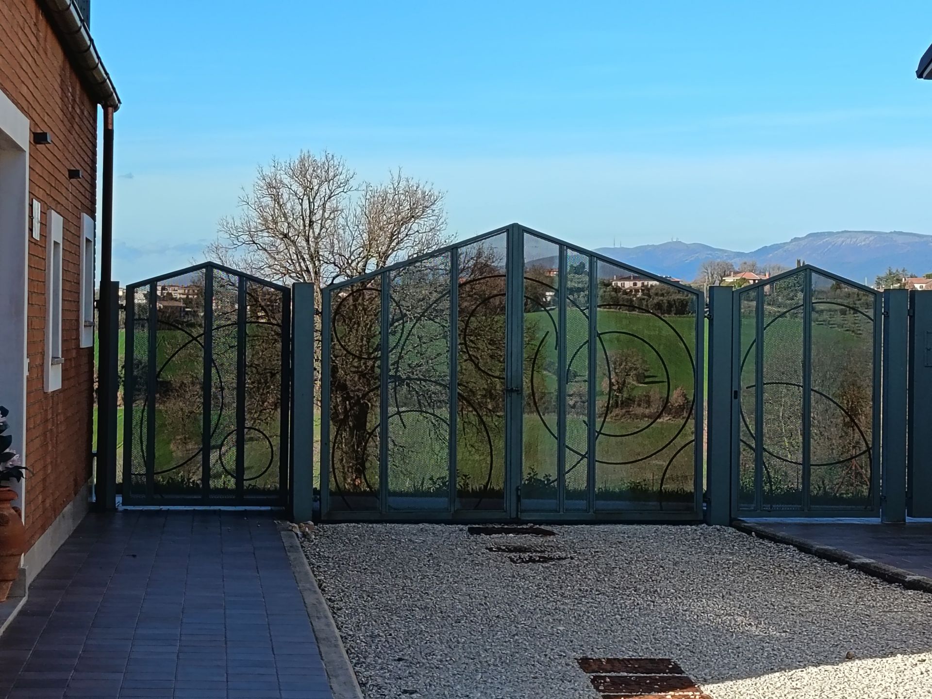 Un cancello di metallo scuro con motivi decorativi a spirale si erge tra un muro di mattoni e un campo