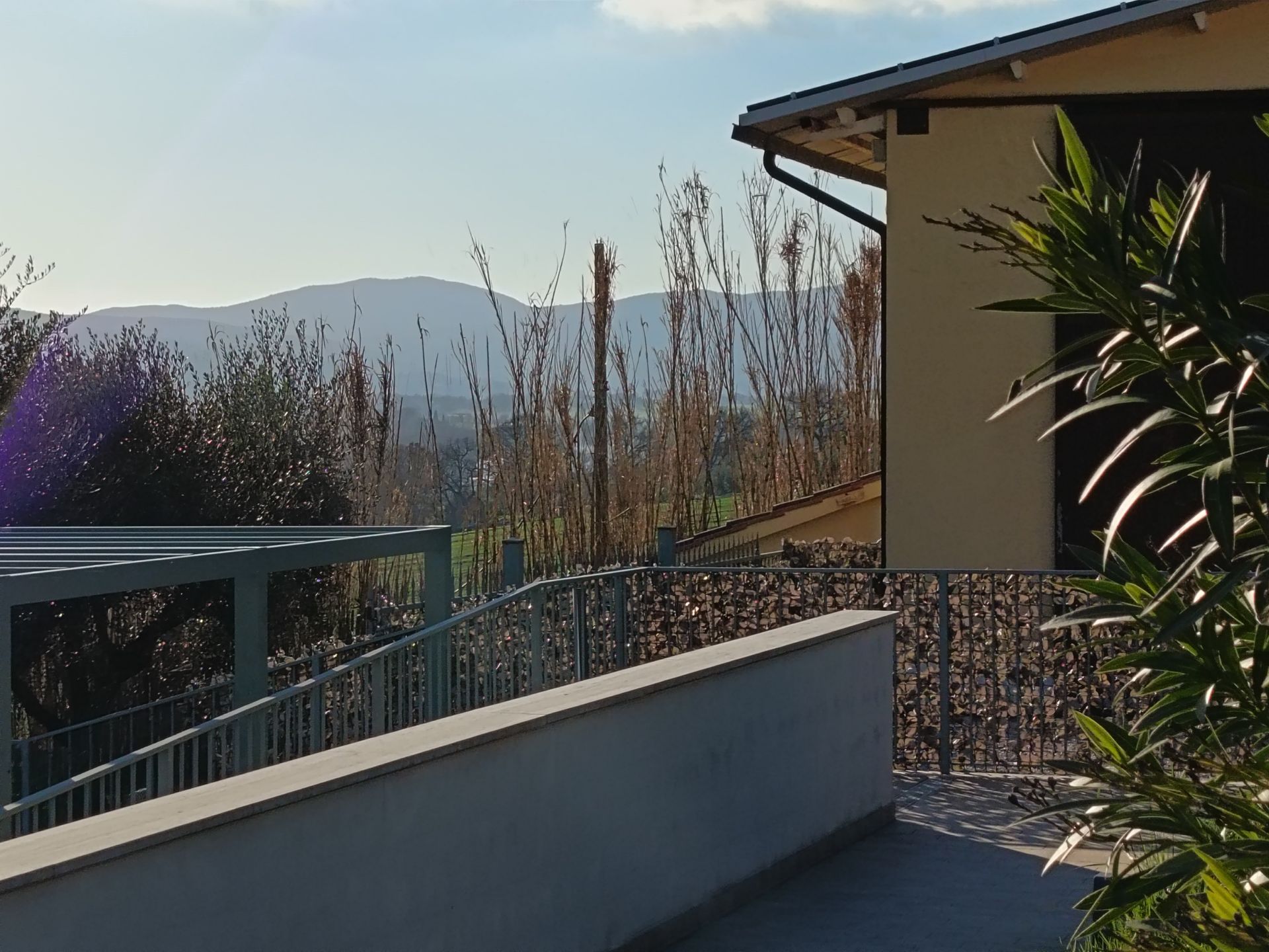 Una terrazza con muro in pietra e ringhiera in metallo si affaccia su un paesaggio collinare alberato sotto un cielo azzurro