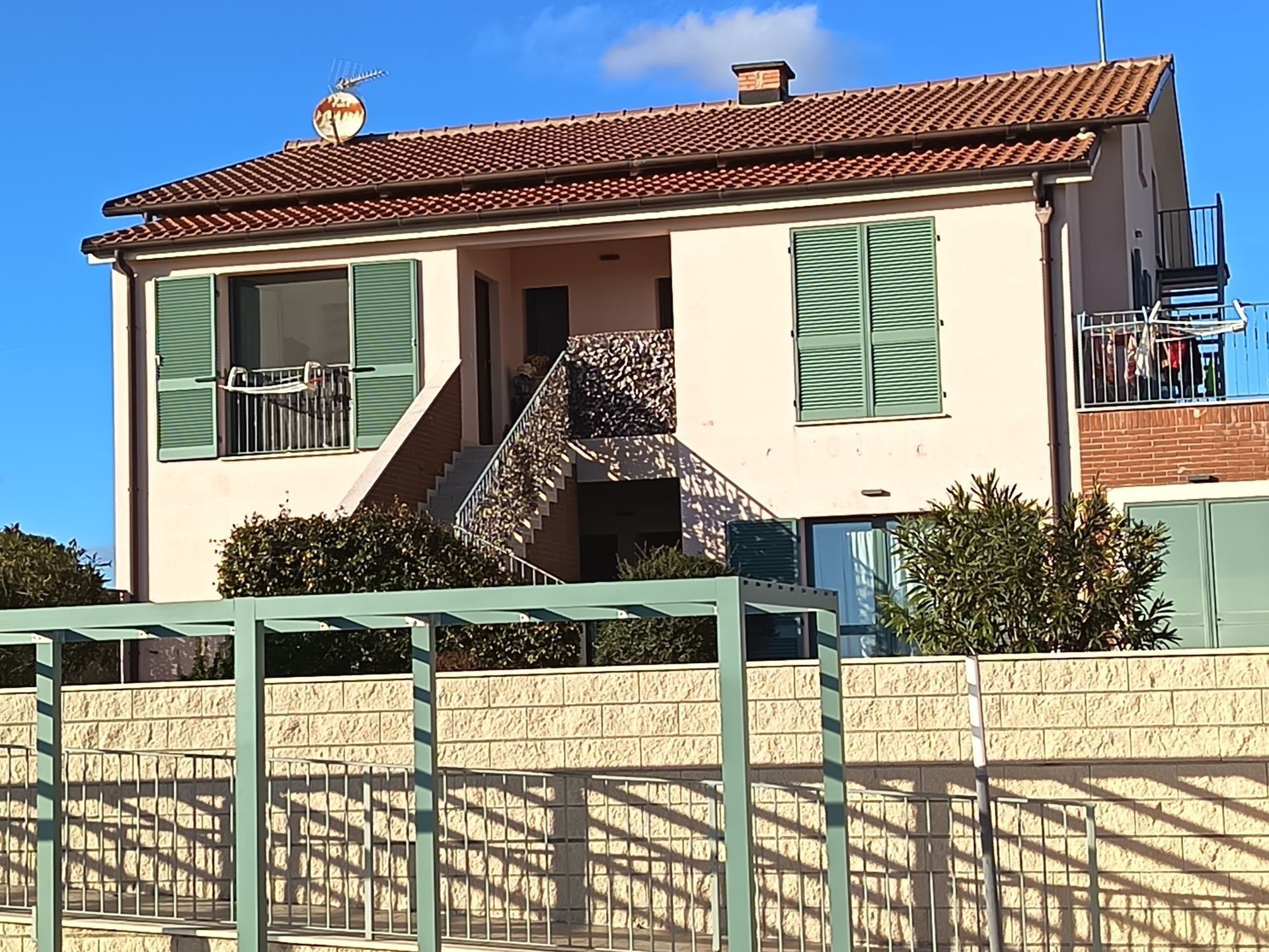 Una casa chiara a due piani con persiane verdi, una scala esterna centrale e un tetto di tegole sotto un cielo azzurro