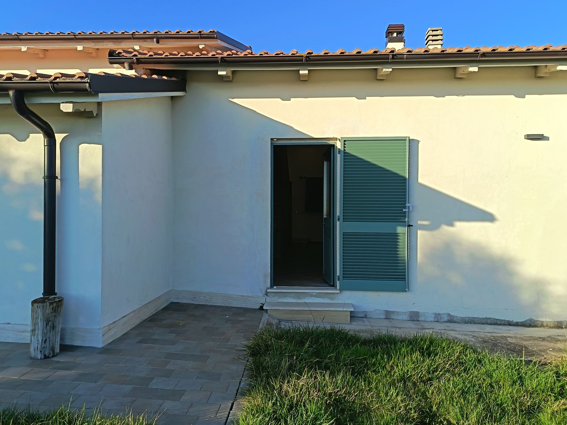 Esterno di una casa dai colori chiari con un'unica porta aperta, persiane verdi, un pluviale e un patio pavimentato.