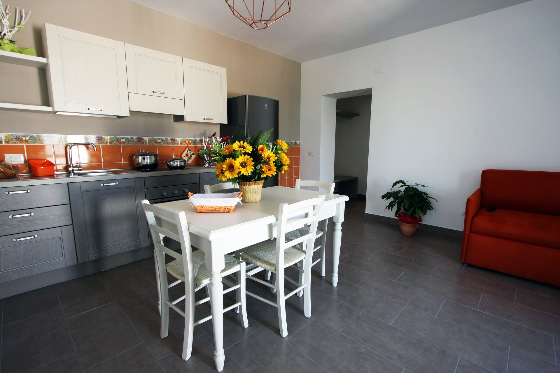 Una cucina e una zona giorno con tavolo e sedie da pranzo bianchi, mobili grigi, girasoli e un divano arancione.