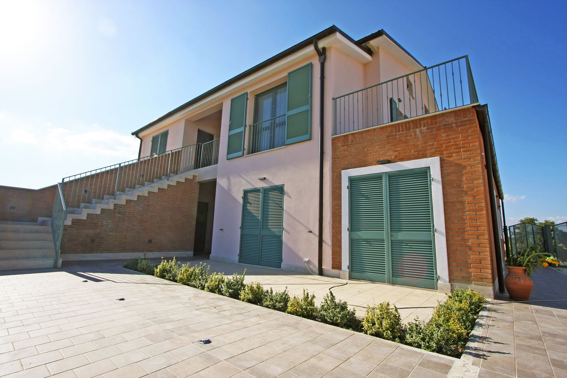 Una villa a due piani con pareti rosa e in mattoni, persiane verdi e una scala esterna che conduce a una terrazza superiore.