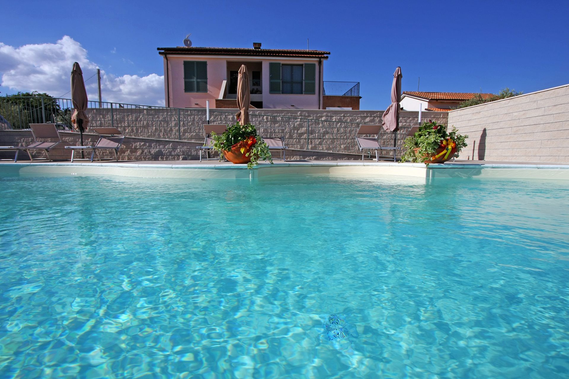 Una piscina turchese brillante di fronte a una casa rosa con un muro di contenimento in pietra e lettini prendisole.