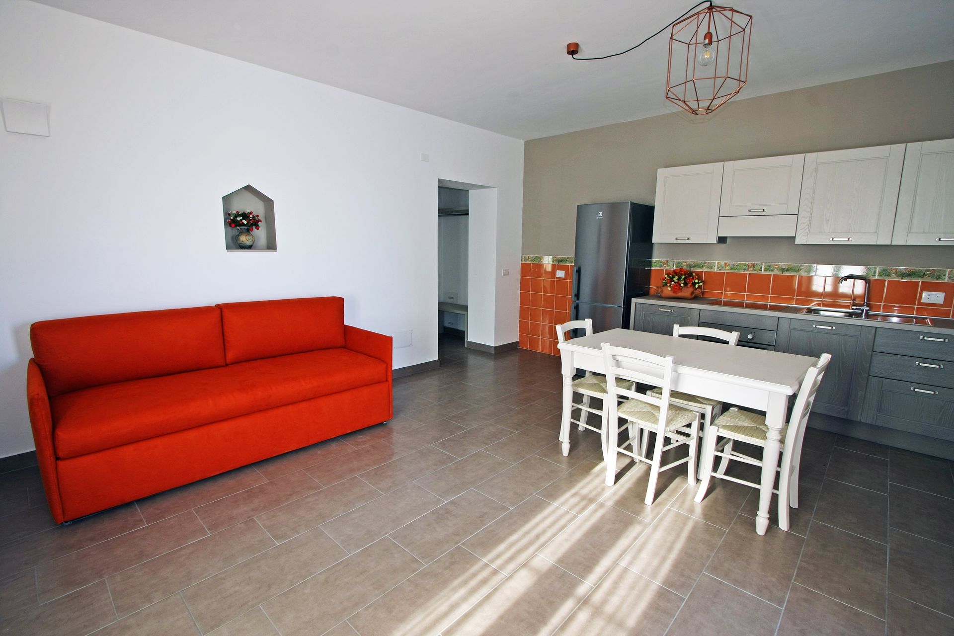 Un luminoso soggiorno con divano rosso, tavolo da pranzo con sedie e cucina con piastrelle arancioni e mobili grigi.