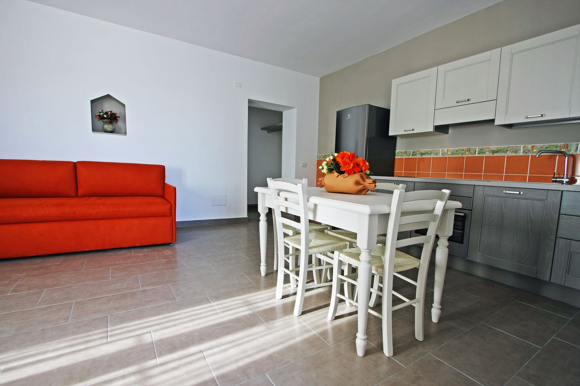 Un open space con divano arancione, tavolo da pranzo bianco con sedie e cucina grigia con piastrelle arancioni alle pareti.
