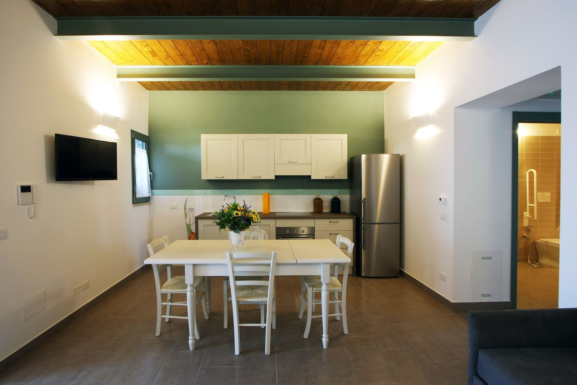 Un monolocale con tavolo da pranzo, mobili da cucina bianchi, frigorifero in acciaio inox e una porta che conduce al bagno.