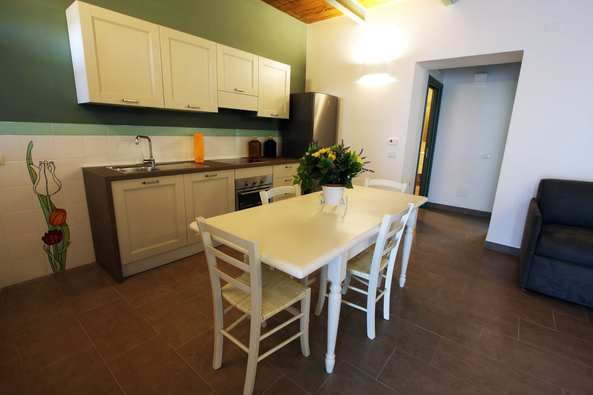 Cucina e zona pranzo con tavolo chiaro, sedie in legno, mobili color crema e pavimento in piastrelle scure.