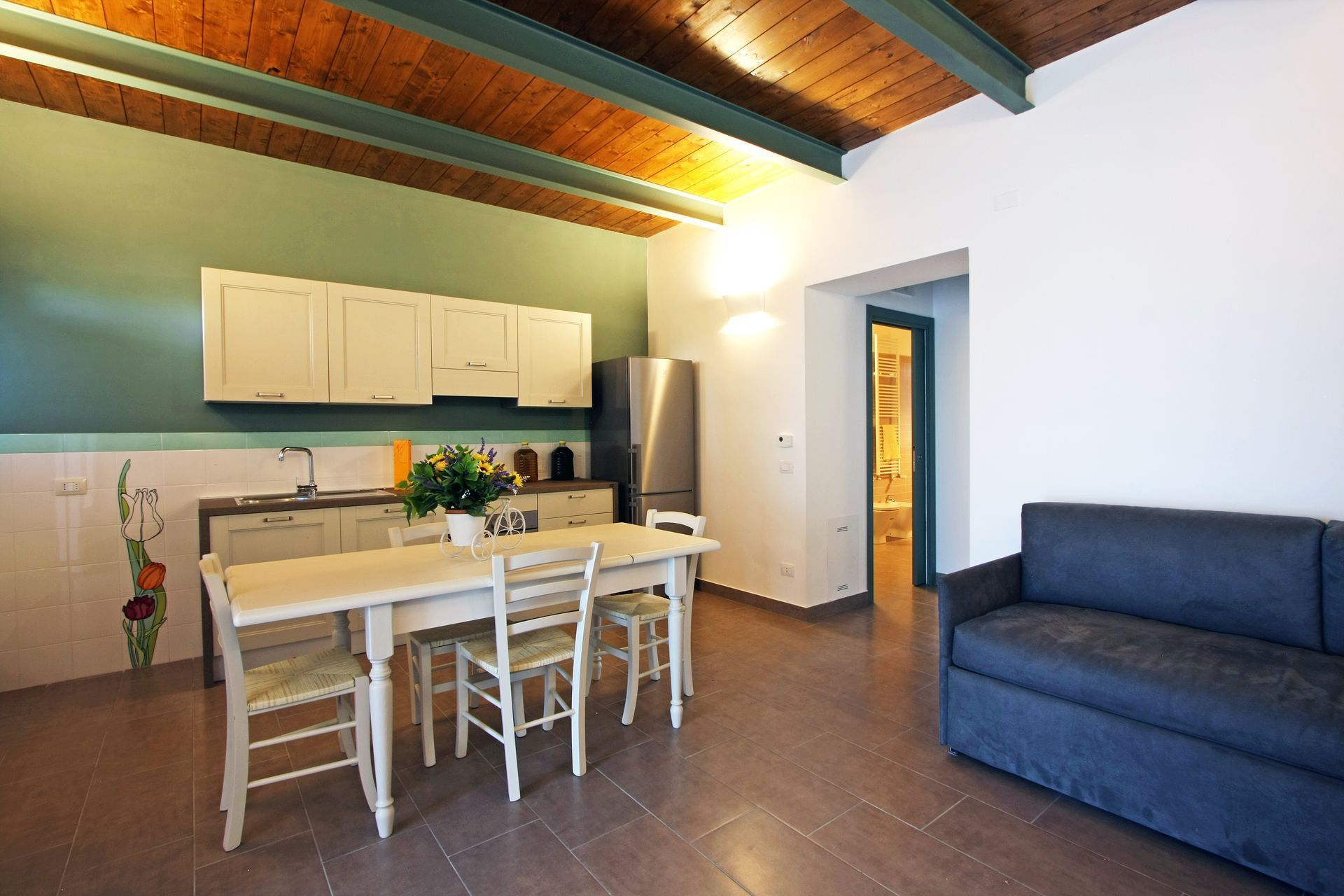 Una cucina-sala da pranzo con tavolo, sedie, armadietti, frigorifero e un divano blu scuro sotto un soffitto in legno.