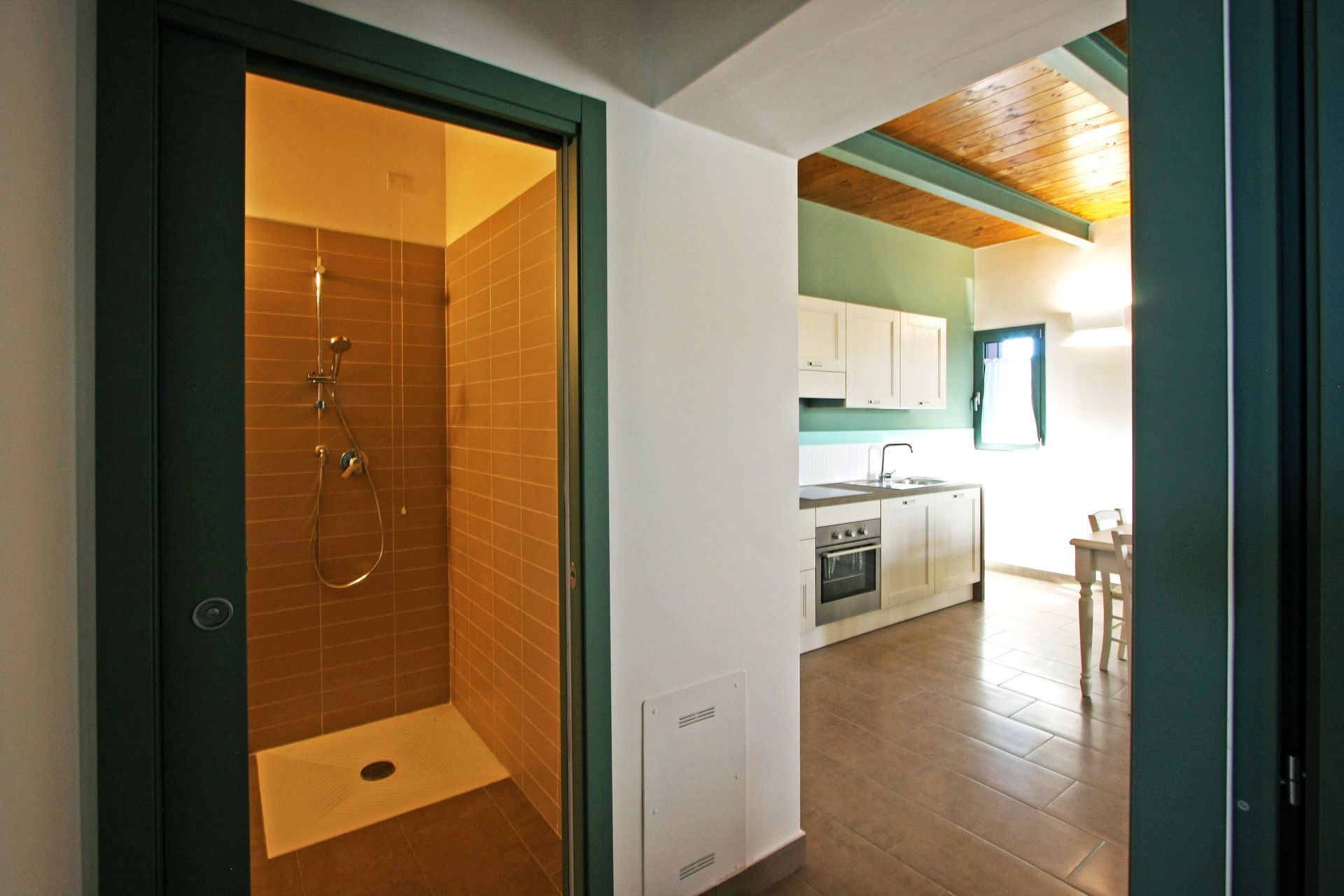 Una vista attraverso una porta su un box doccia piastrellato, con una cucina a vista e una zona pranzo sullo sfondo.