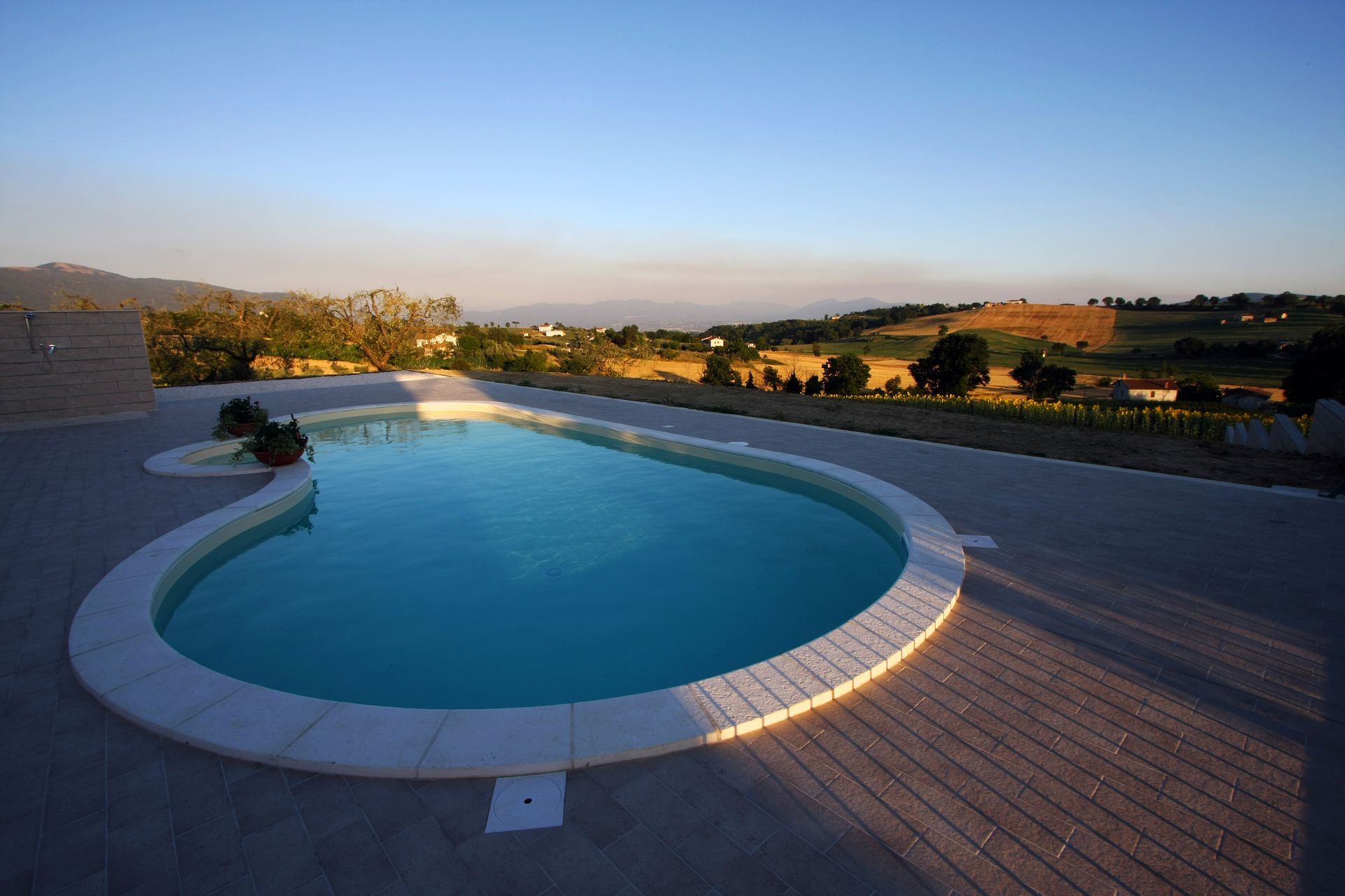 Una piscina ovale color turchese, situata su un patio in pietra con vista su un paesaggio rurale al crepuscolo.