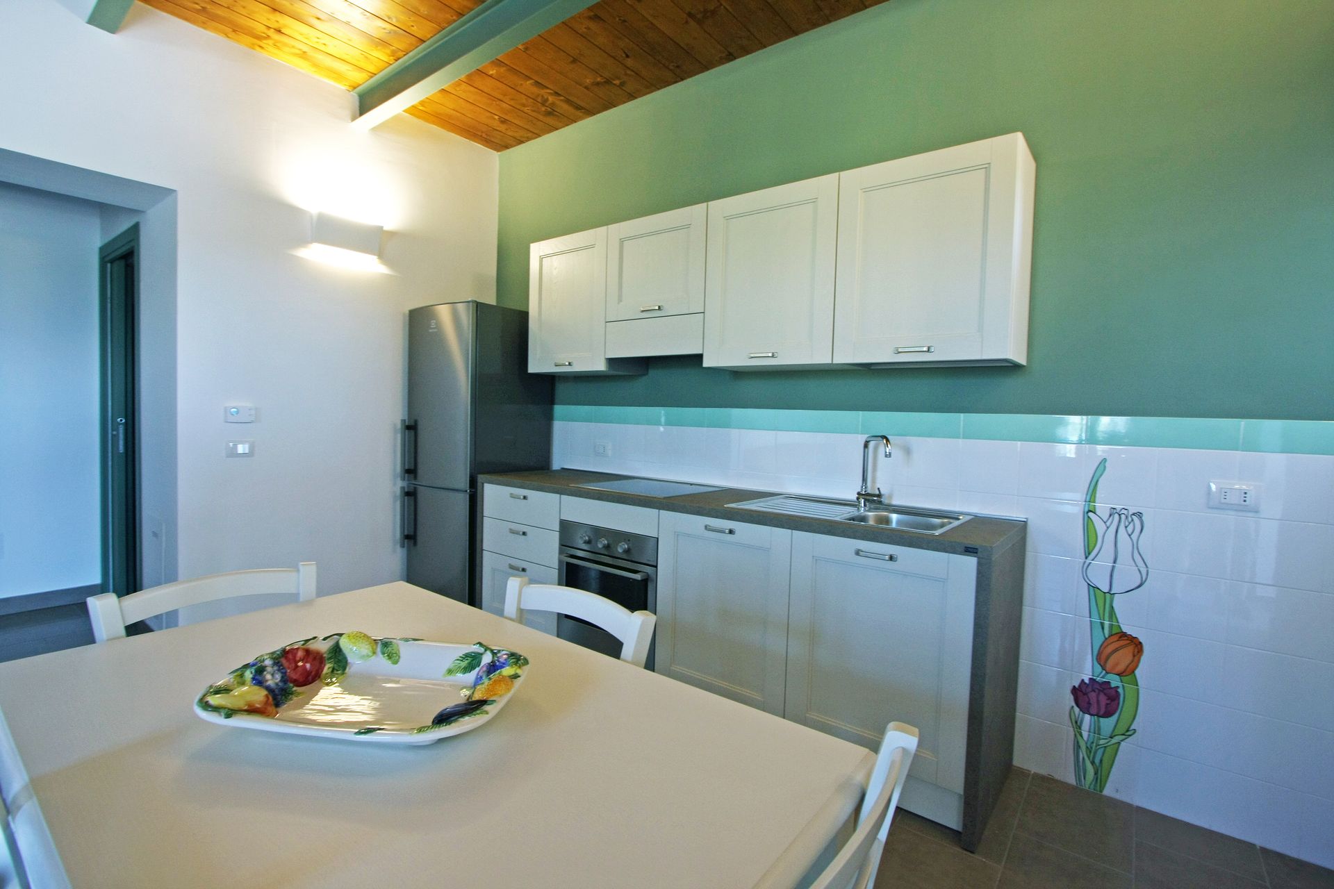 Una cucina con mobili bianchi, un frigorifero in acciaio inox e un tavolo da pranzo, addossata a una parete verde salvia.