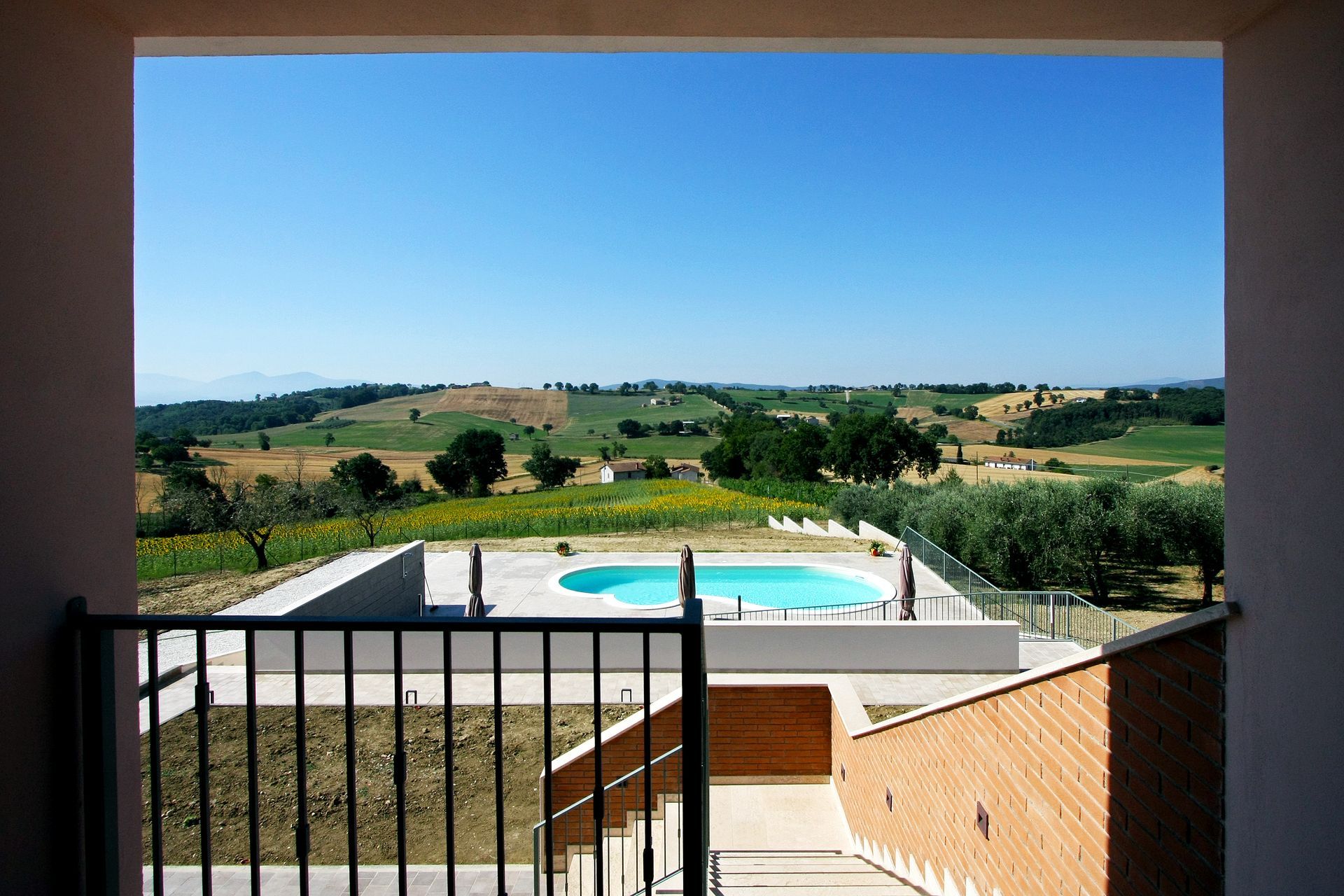 Una vista panoramica da un balcone che si affaccia su una piscina, prati e dolci colline sotto un cielo azzurro brillante.