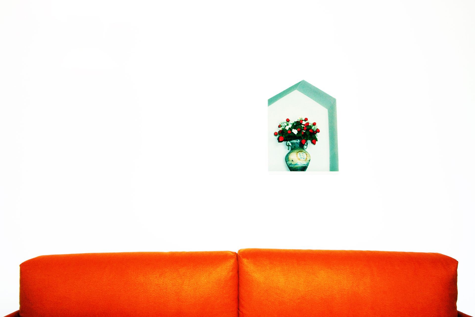 Un divano arancione appoggiato a una parete bianca con una cornice che raffigura dei fiori in un vaso blu.