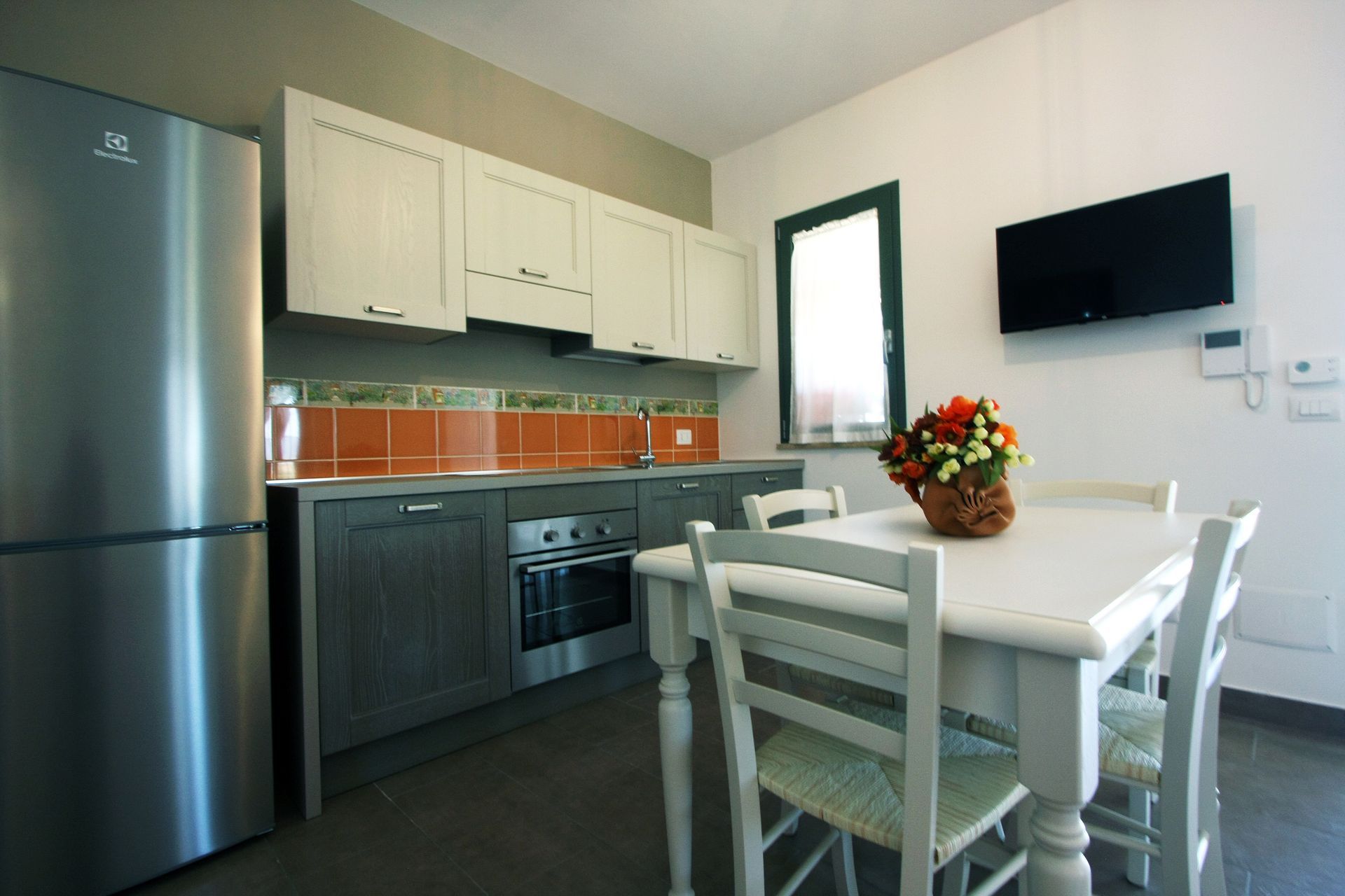 Una cucina con frigorifero in acciaio inox, pensili bianchi, mobili base grigi, tavolo da pranzo e televisore a parete.