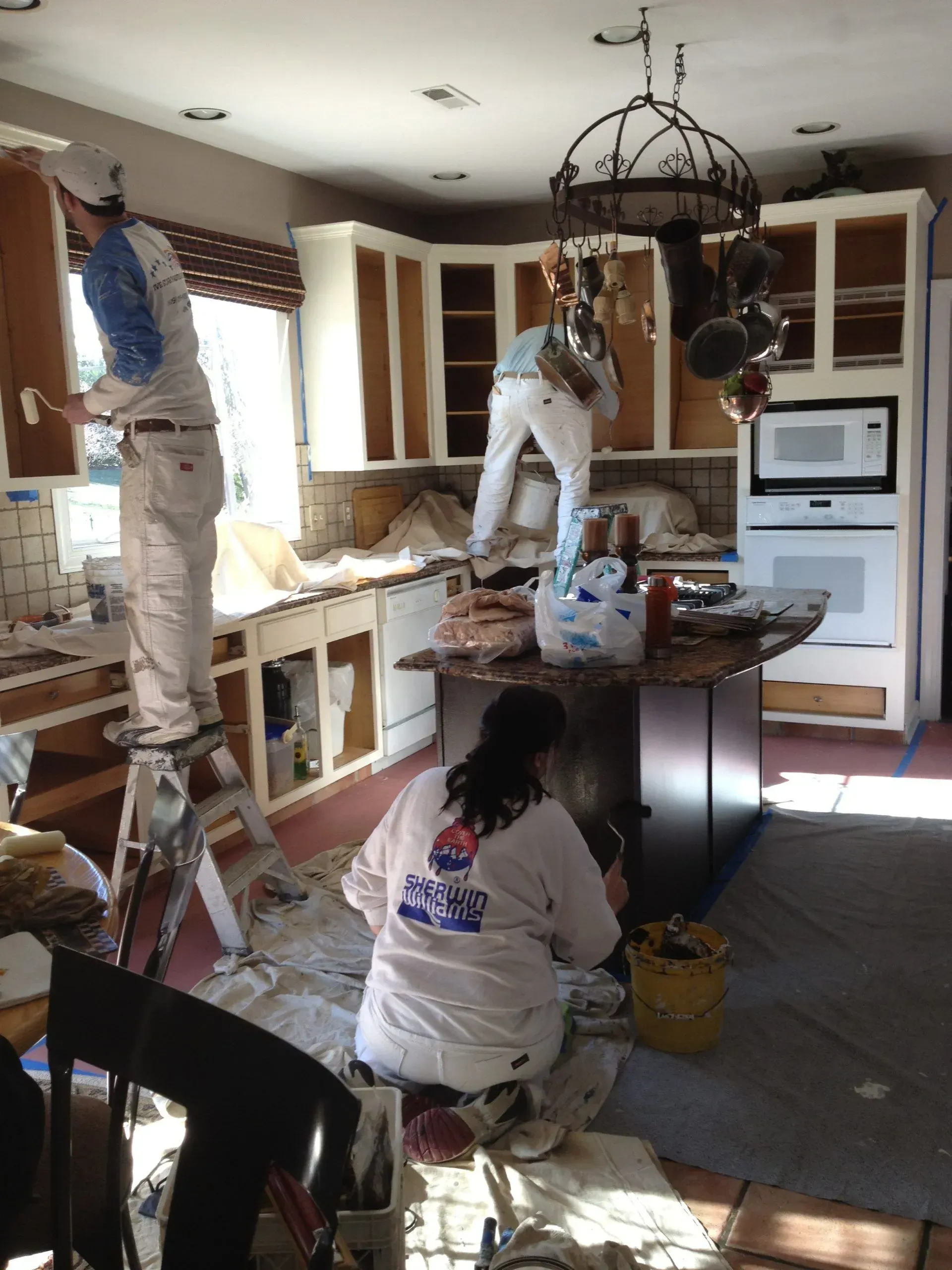 A woman is kneeling down in a kitchen while two men paint the cabinets