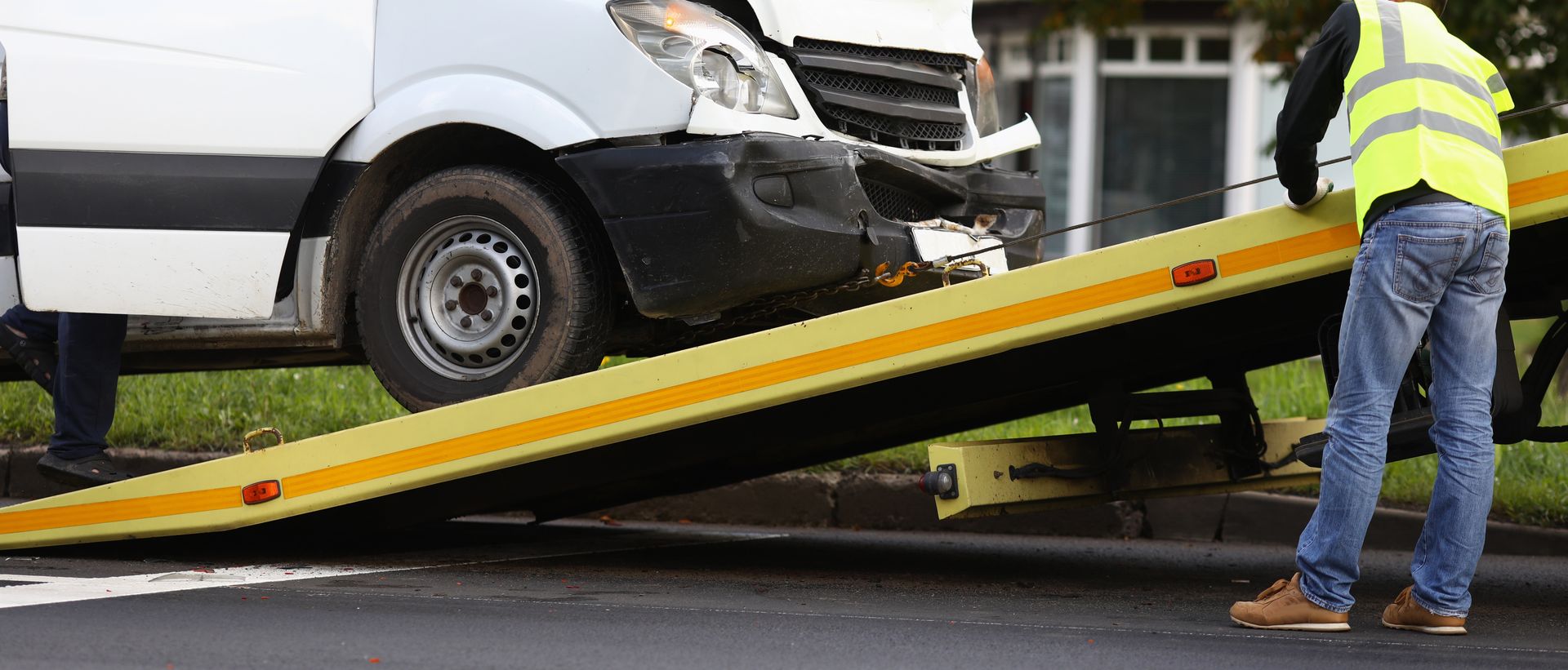 Damaged white van being winched onto a tow truck ramp by a worker in a safety vest. Damaged white van being winched onto a tow truck ramp by a worker in a safety vest.