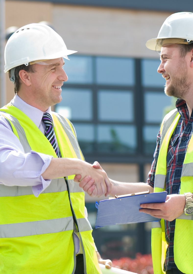 Two construction workers are shaking hands in front of a building.