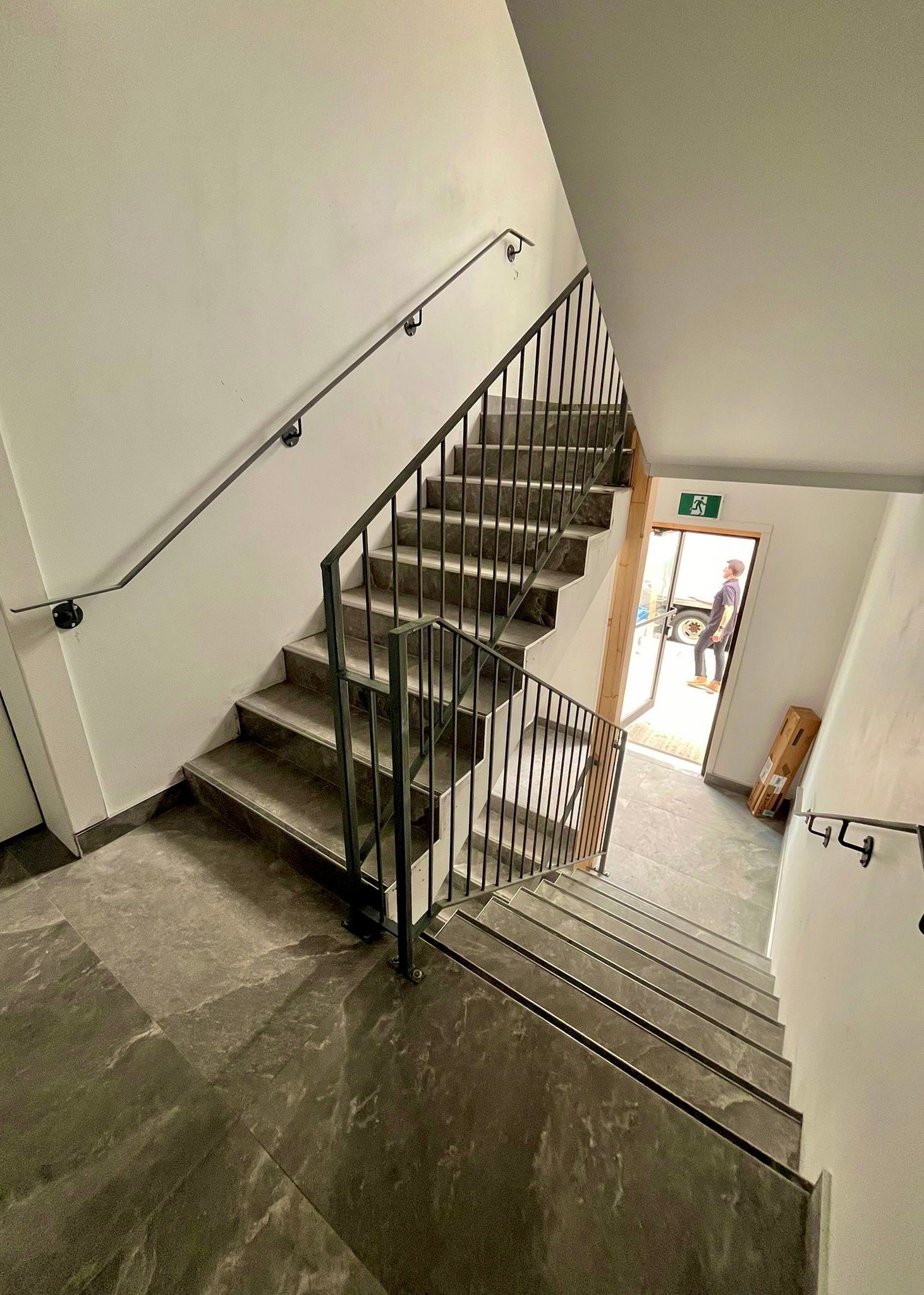 Un escalier descend de l'étage. Marches et murs gris, rampes noires et porte ouverte au bout.