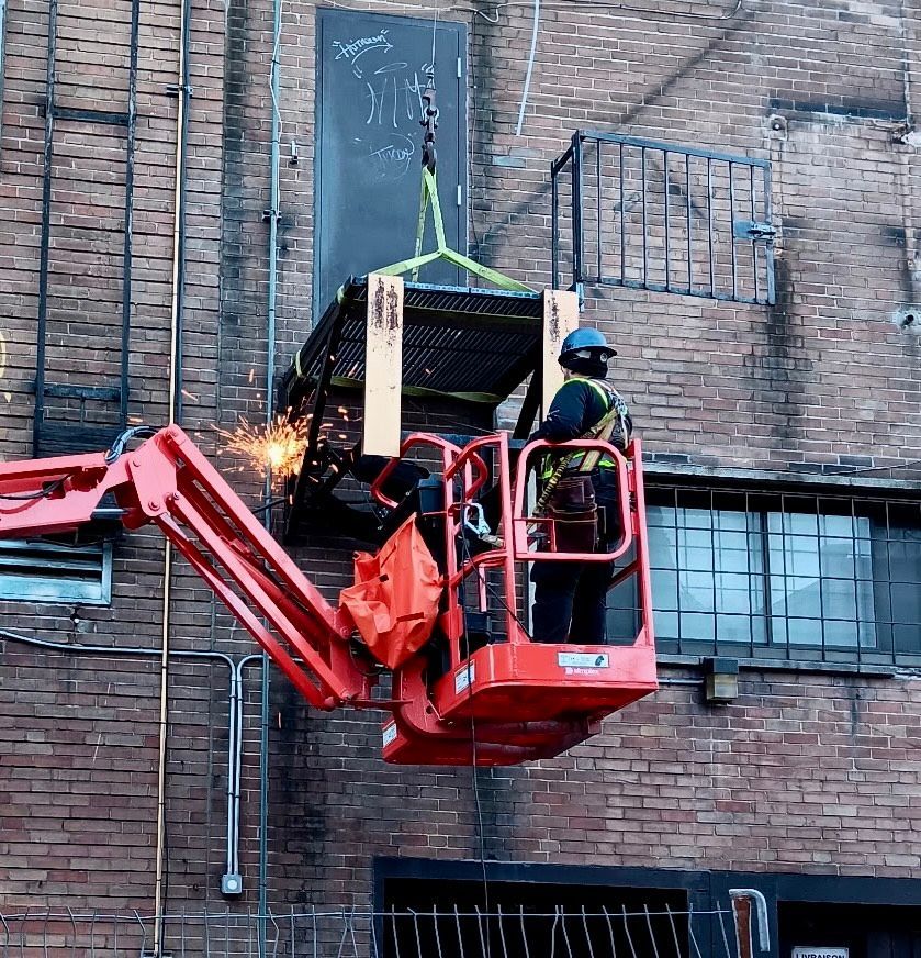 A construction worker in a lift welding a metal cage attached to a crane, outside a brick building.
