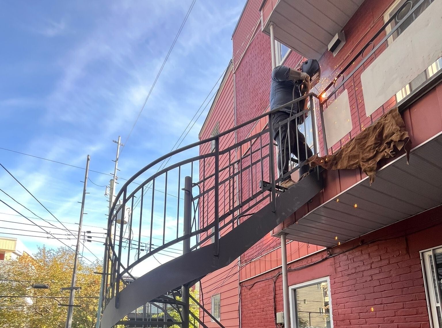 Un soudeur travaille sur un escalier métallique fixé à un bâtiment en briques rouges, sur fond de ciel bleu.