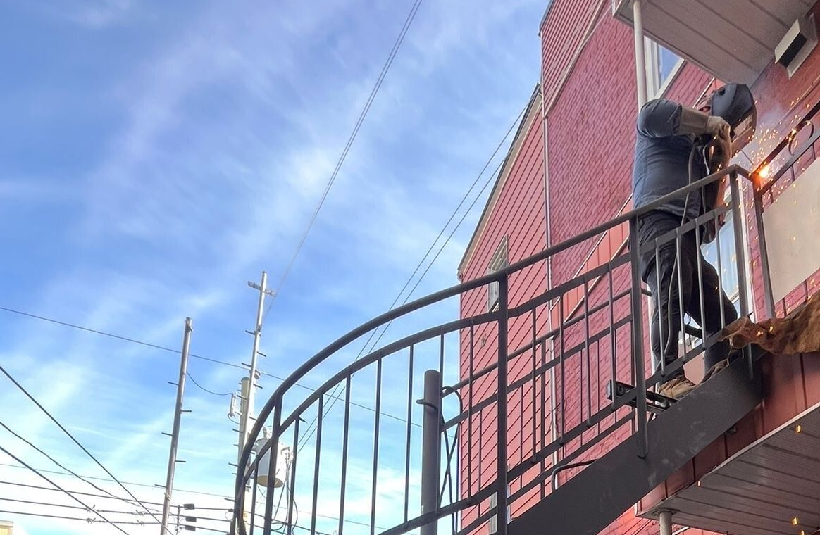 Un soudeur travaille sur un escalier en colimaçon extérieur, à côté d'un bâtiment en briques rouges, sous un ciel bleu.