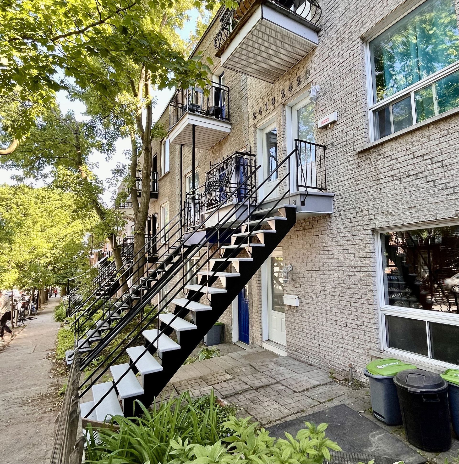 Exterior view of a brick building with exterior stairs leading to upper-floor entrances. Street and trees in the background.