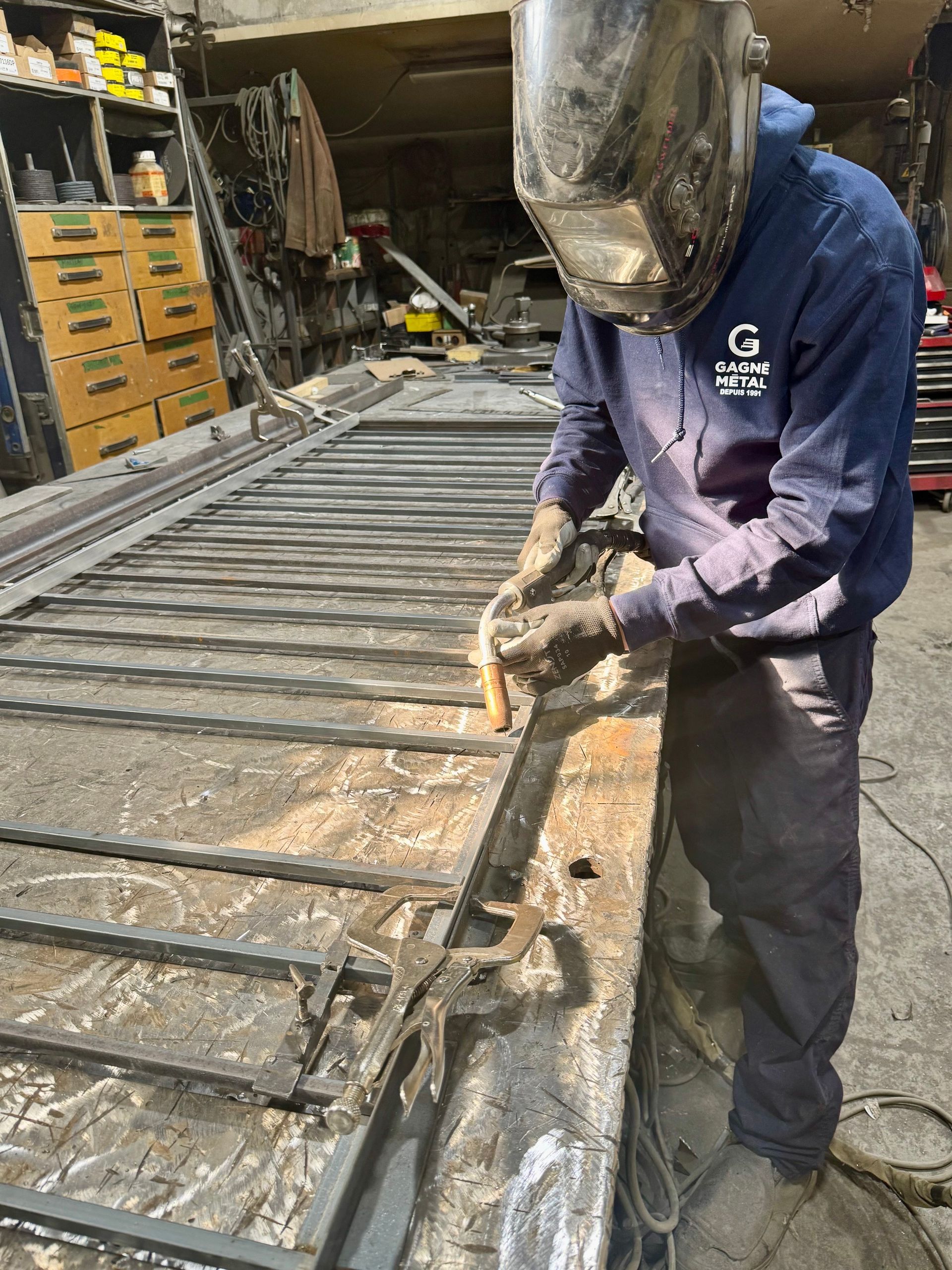 Welder in a workshop, wearing a helmet and gloves, welding metal frame.
