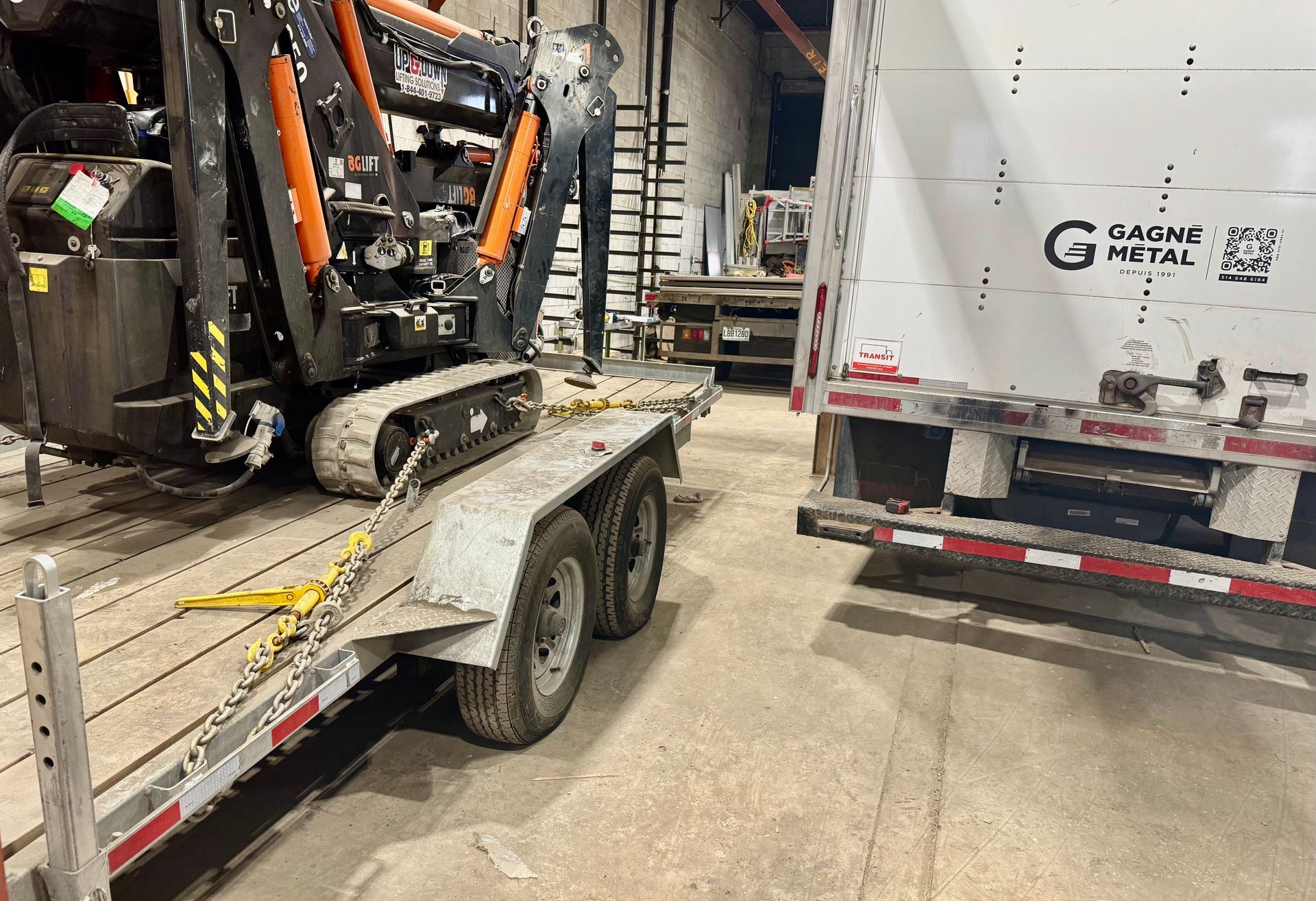 Mini excavator secured on a trailer in a warehouse. A large white structure is beside the trailer.