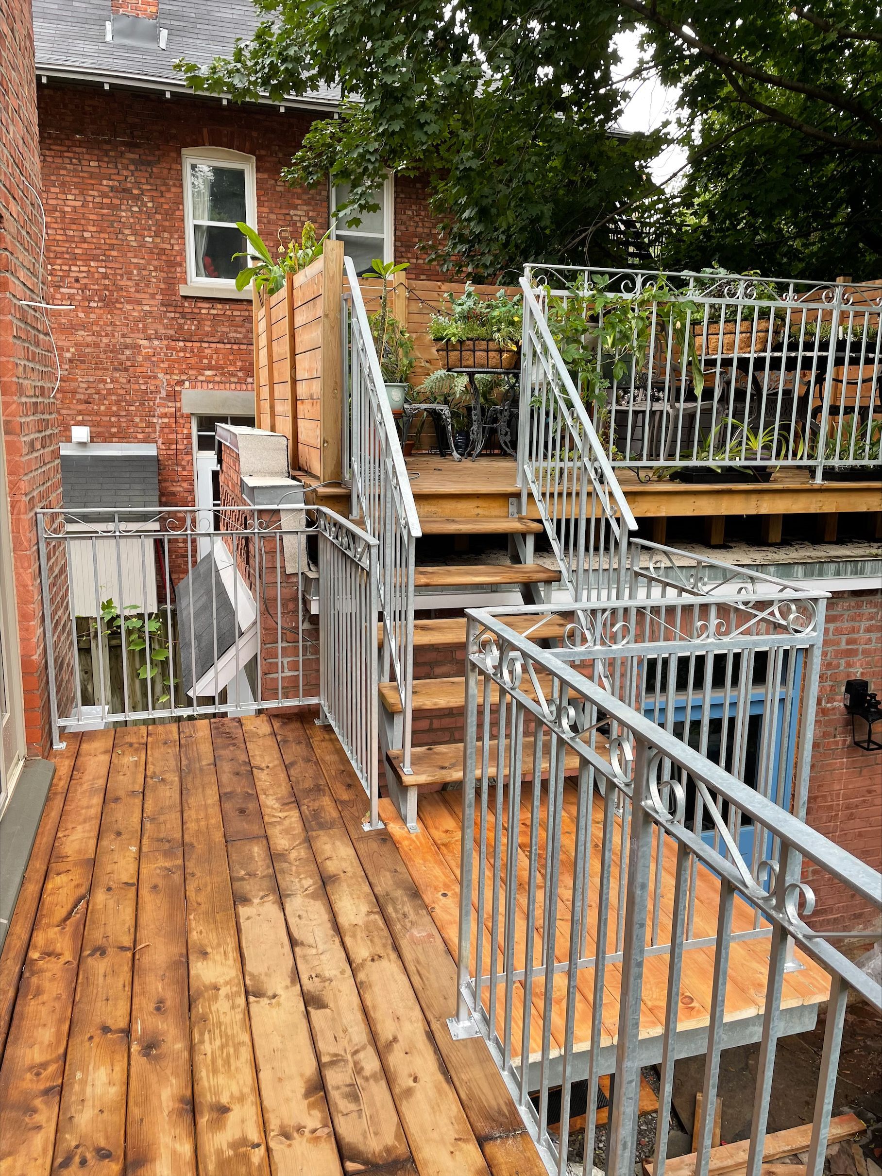 Une terrasse en bois avec des escaliers et une rampe métallique devant un bâtiment en brique.