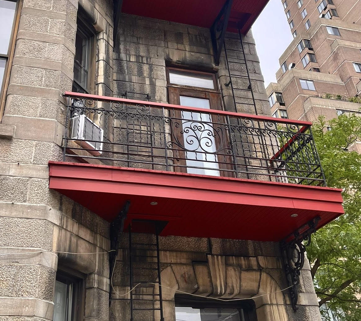 Balcon avec garniture rouge et garde-corps en métal noir sur un bâtiment en pierre.