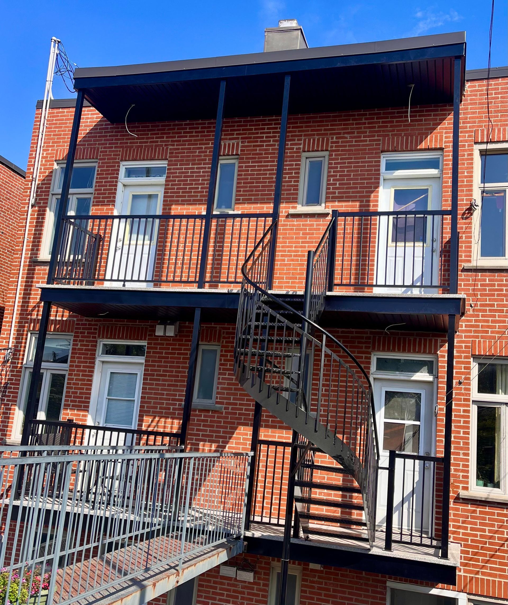 Bâtiment en briques avec balcons en métal noir et escalier en colimaçon. Journée ensoleillée sous un ciel bleu.