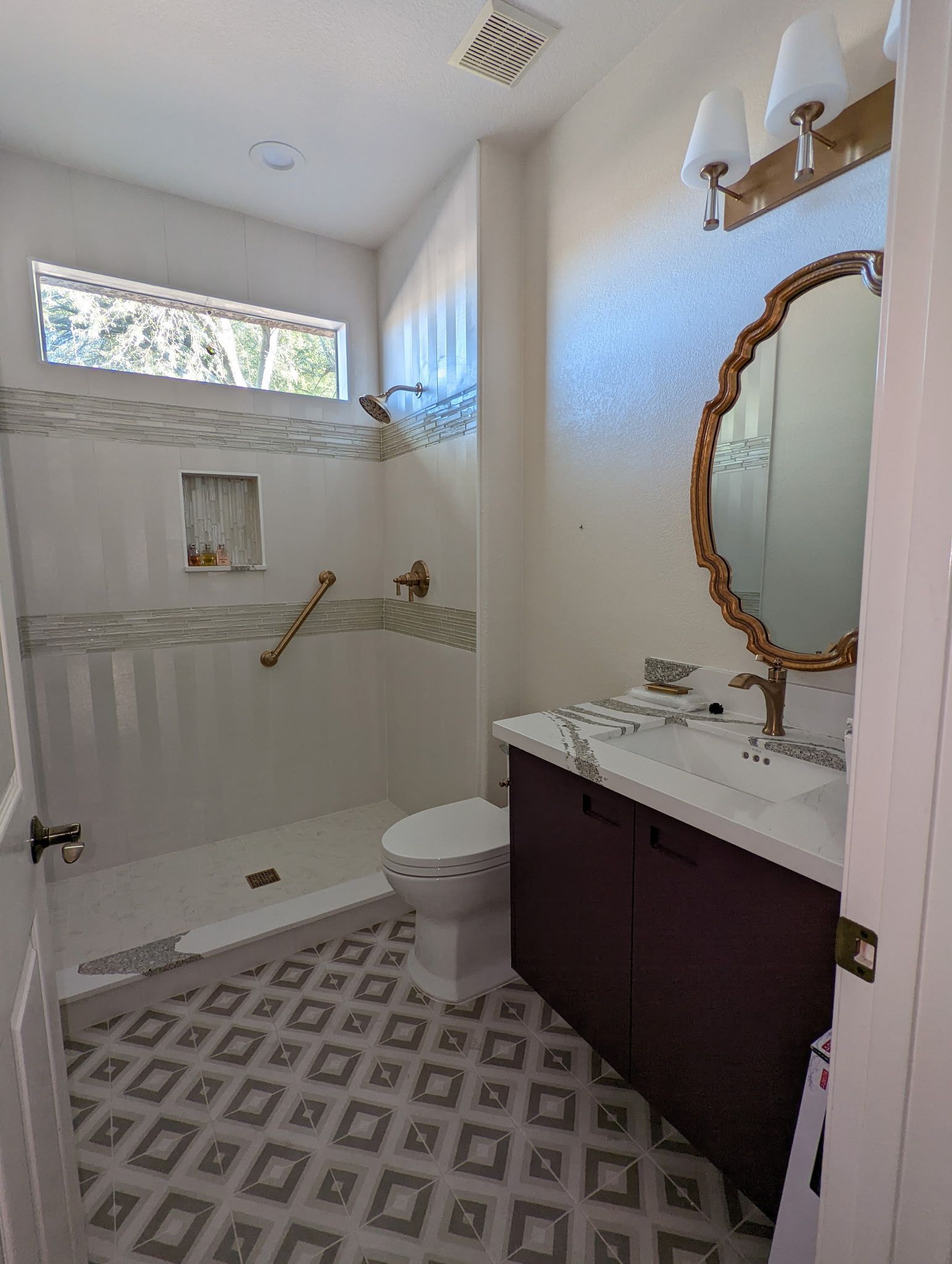 Bathroom with white, gray tile floor, shower, toilet, dark vanity, gold mirror and fixtures.