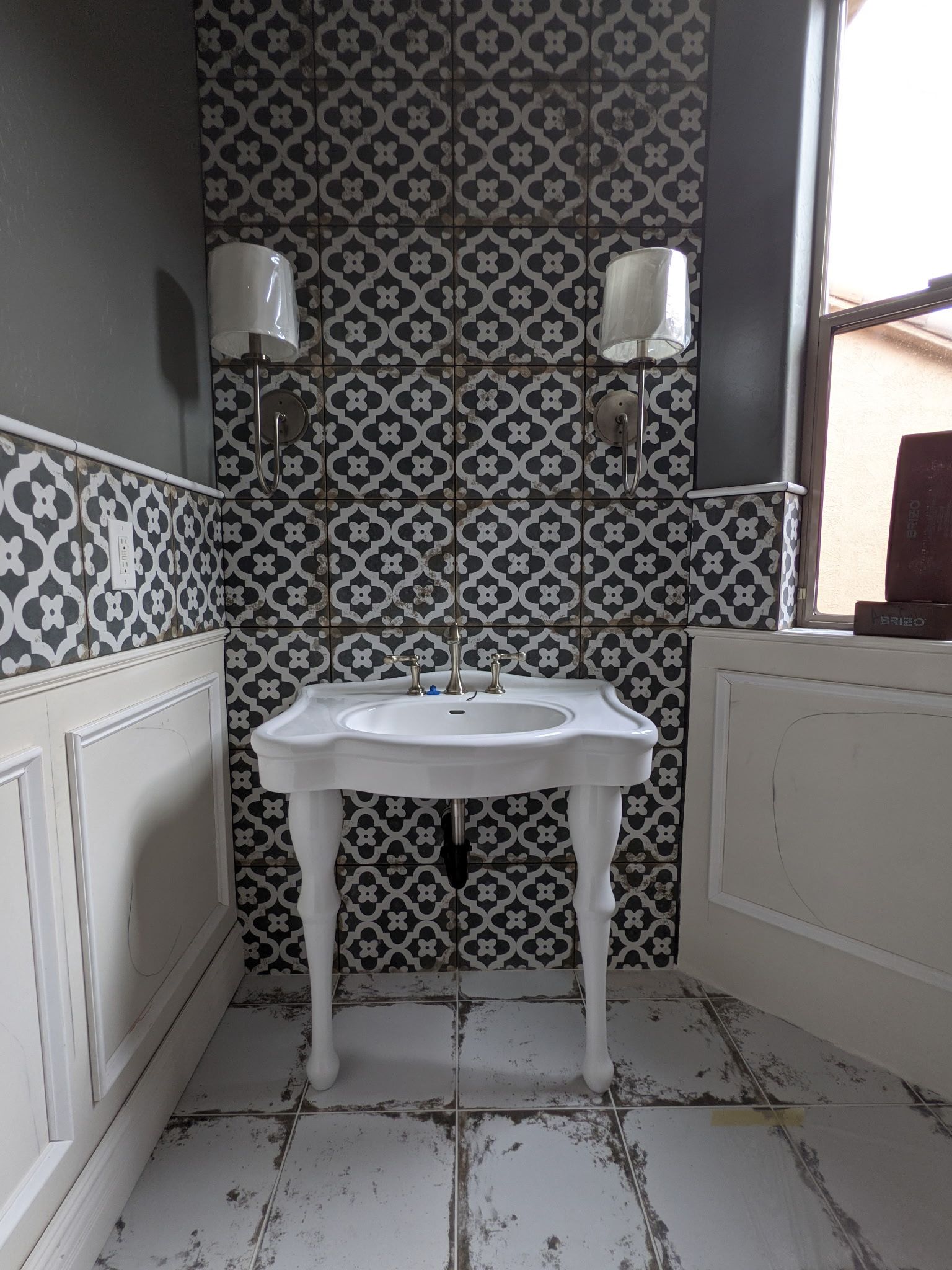 Bathroom with patterned tile accent wall, white pedestal sink, sconces, and gray walls.