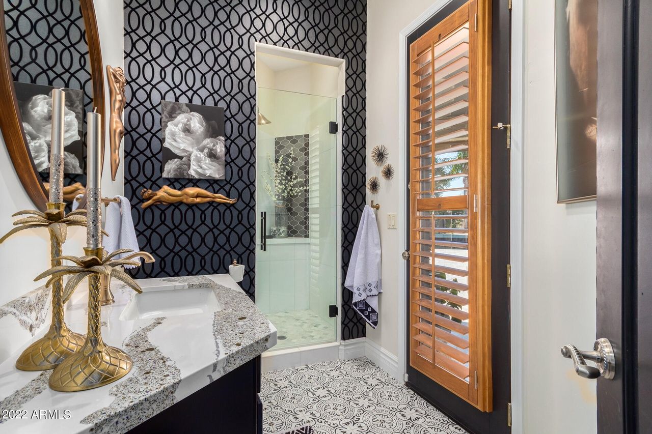 Powder room with patterned wallpaper, marble countertop, and wood-slatted door.