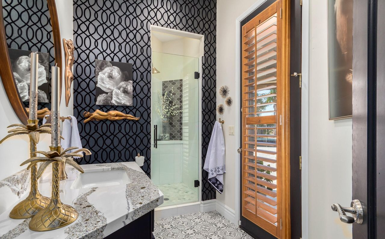 Bathroom with patterned dark wallpaper, glass shower, wooden shutters, and ornate gold accents.