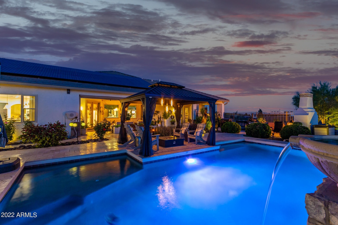Evening view of a pool with water feature, gazebo with seating, and house under a colorful sky.