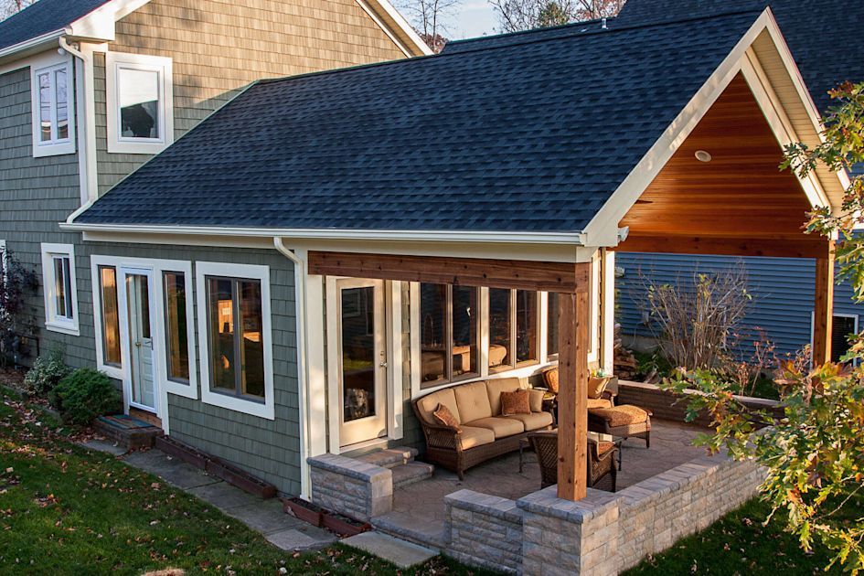 Green house with a sunroom addition, tan seating area, and a brown beam supported overhang.