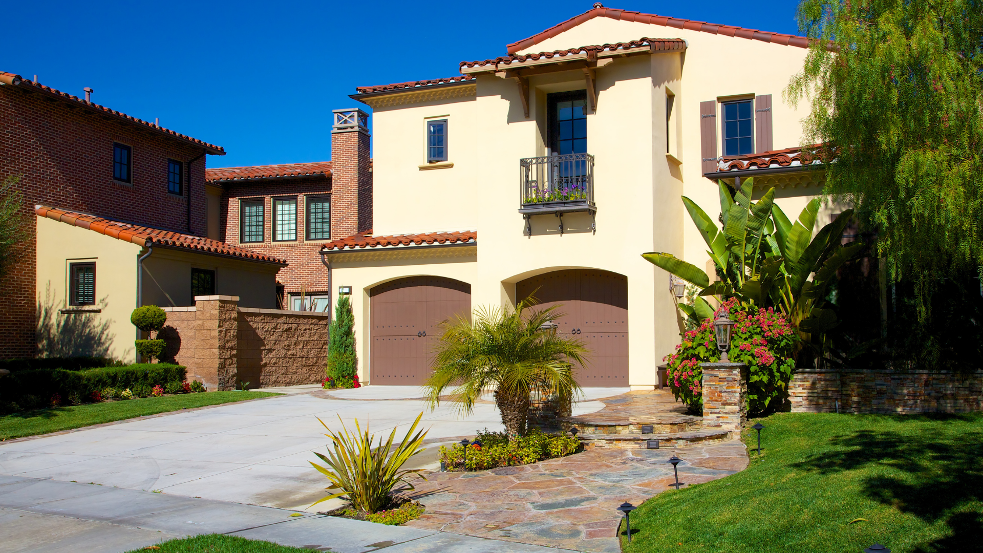 A two-story, cream-colored stucco home with a terracotta tile roof, two-car garage, and a lush front yard.